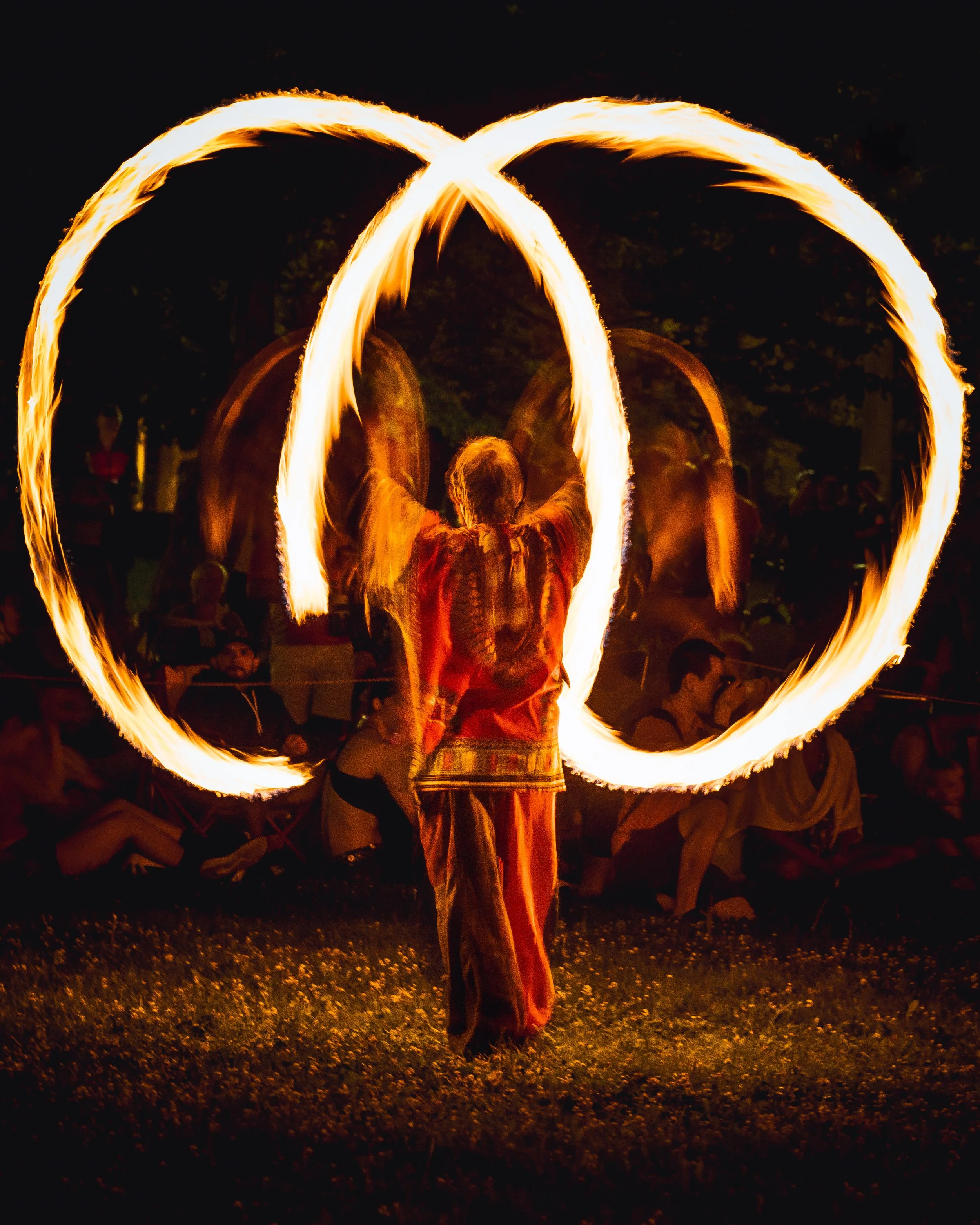 Person performing a fire spinning dance at night, creating large fiery shapes in the air with a fire staff, with an audience sitting in the background.