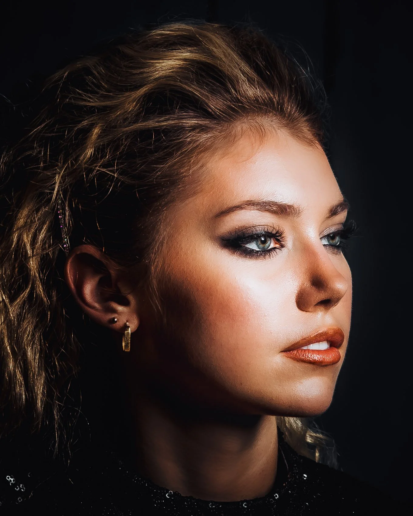 Close-up portrait of a woman with styled hair, makeup, earrings, and a black top, against a dark background.