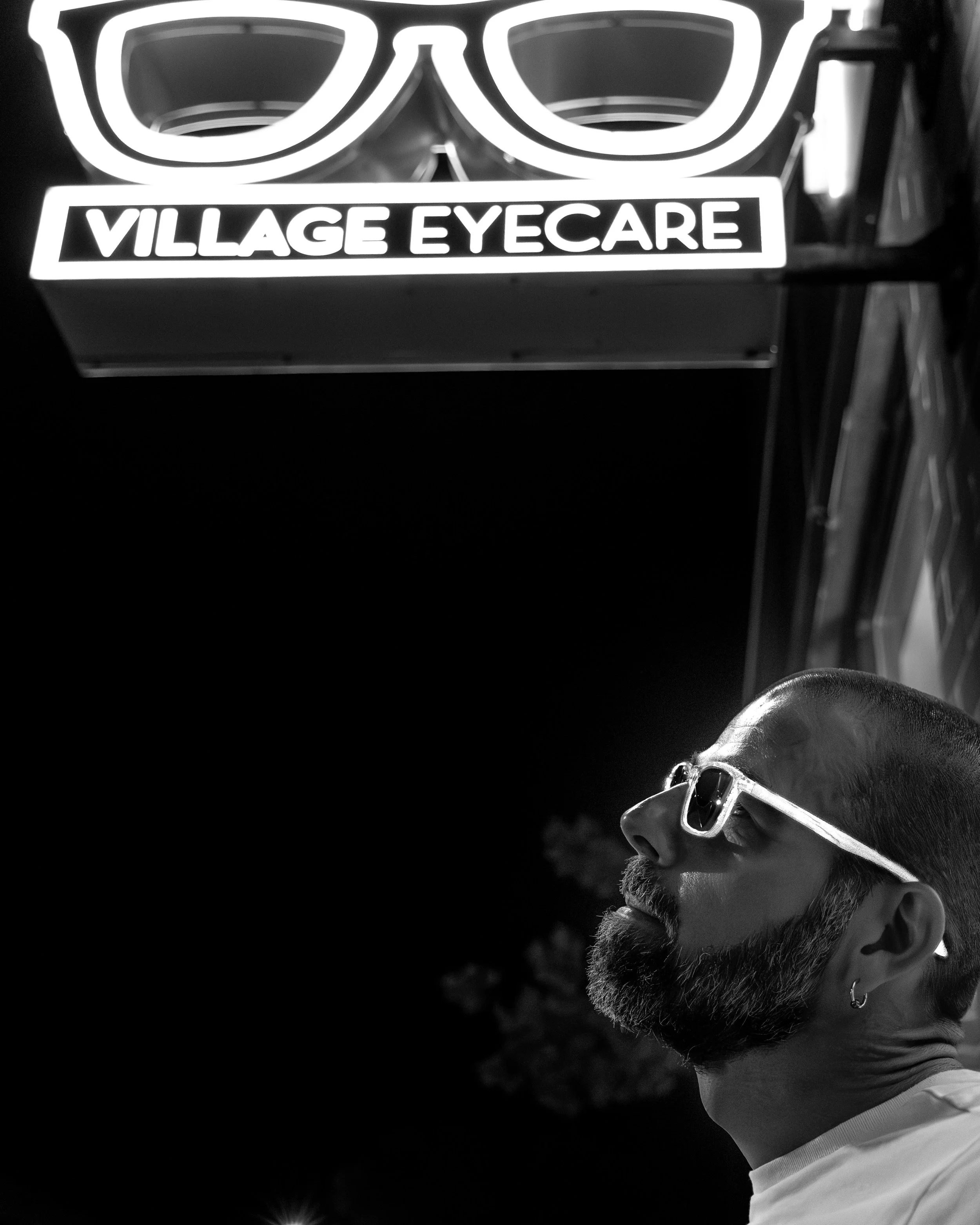 A man with glasses and a beard looking upward at a brightly lit sign that reads "Village Eyecare" with an image of eyeglasses above it, at night.