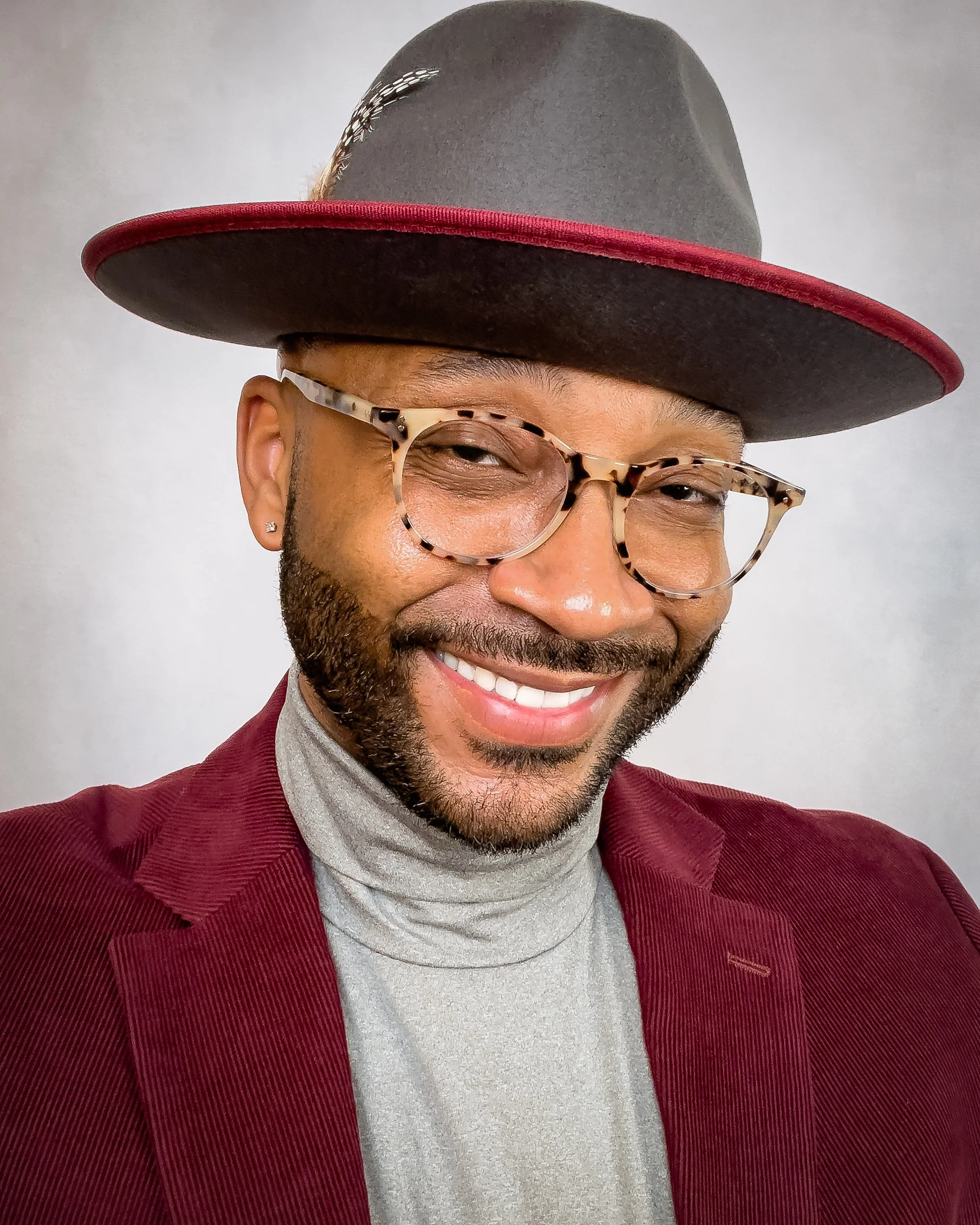 Smiling man wearing tortoiseshell glasses, a gray hat with a red rim, a red blazer, and a light gray turtleneck, standing against a plain background.