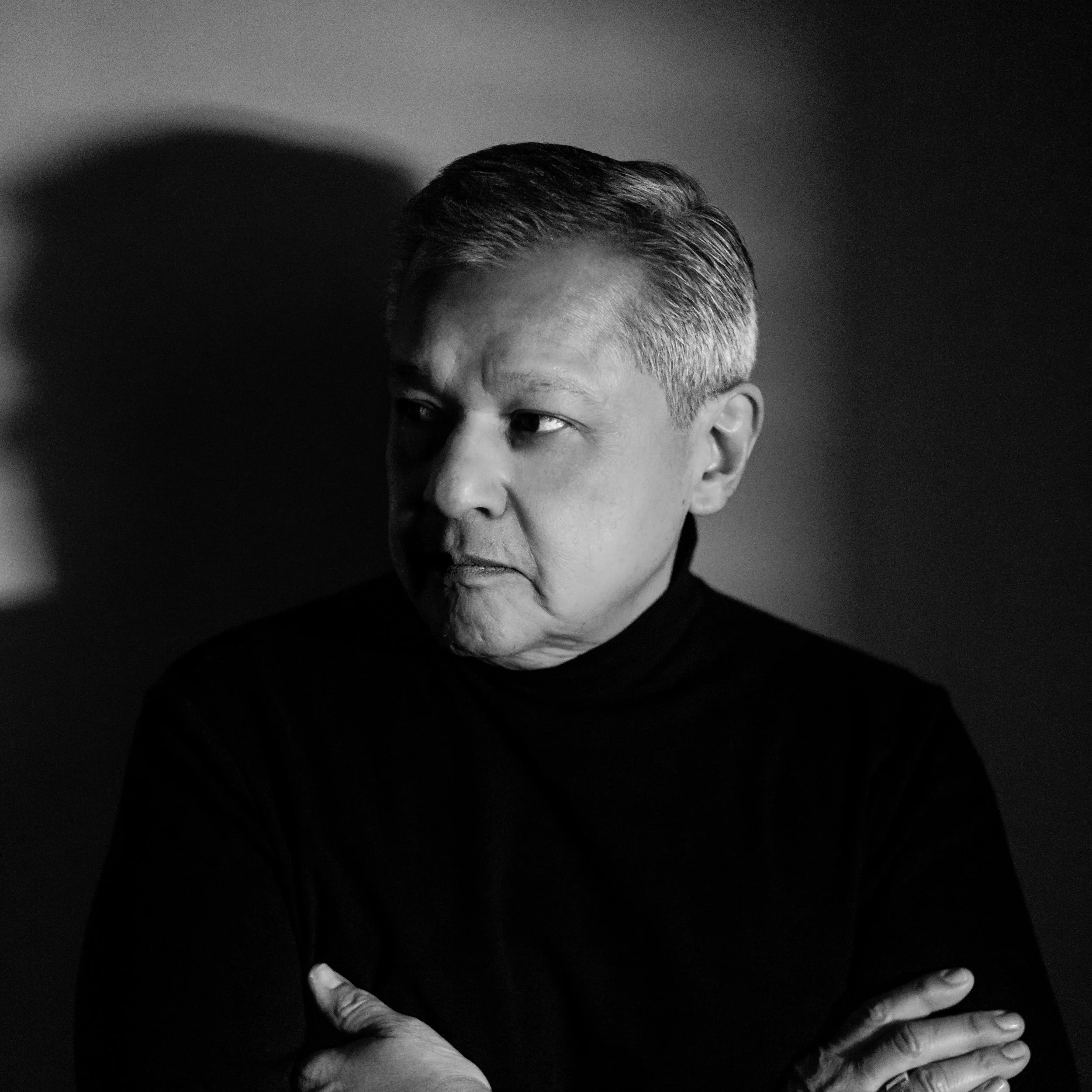 Black and white portrait of a man with short hair, arms crossed, looking to the side in a contemplative pose, wearing a dark turtleneck.