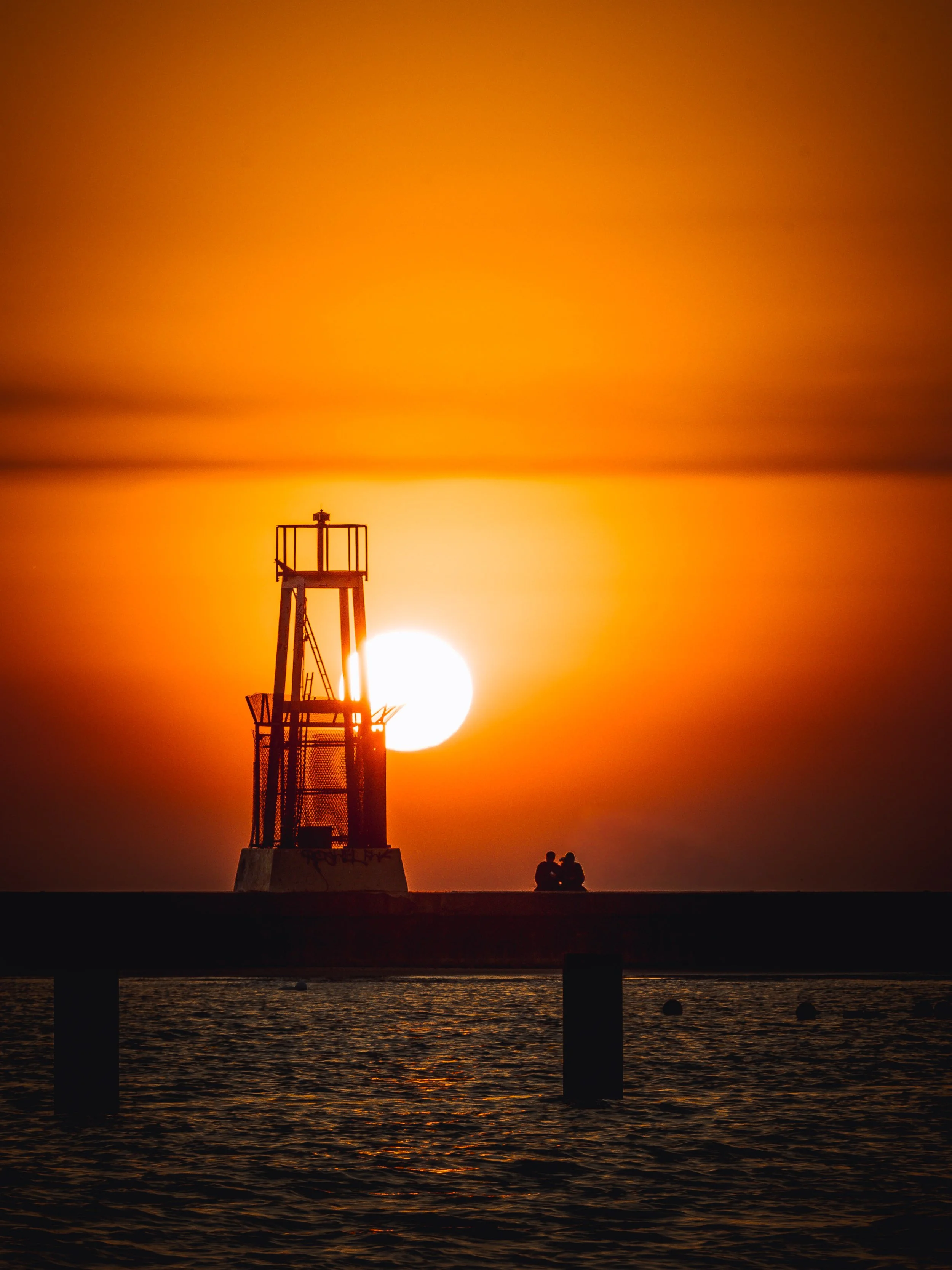 Sunrise over water with a lighthouse silhouette and a couple sitting on a dock.