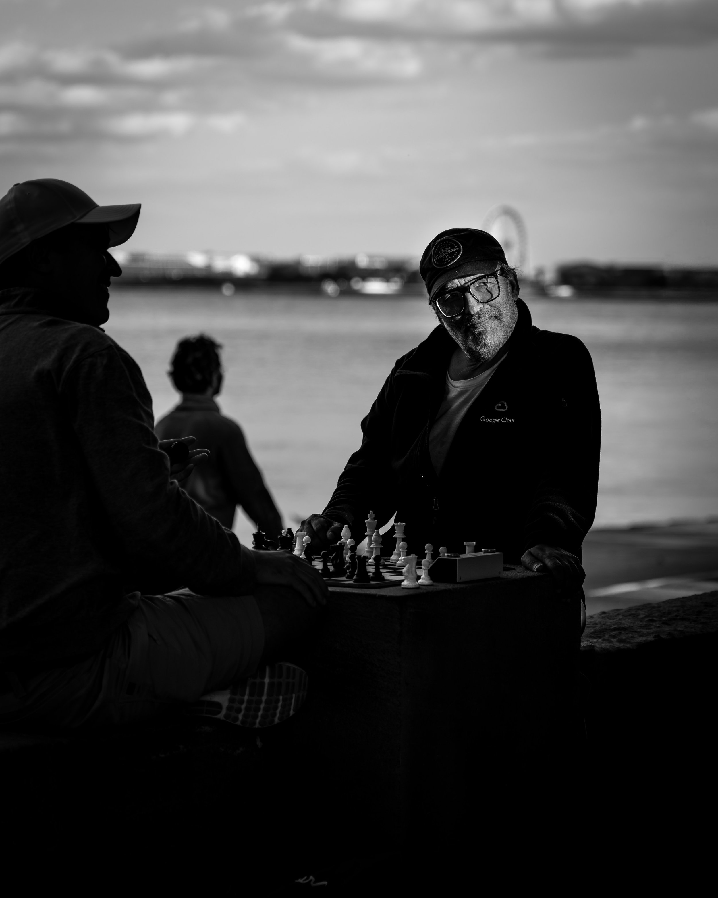 Silhouettes of three men playing chess outdoors near a body of water, with a ferris wheel in the background