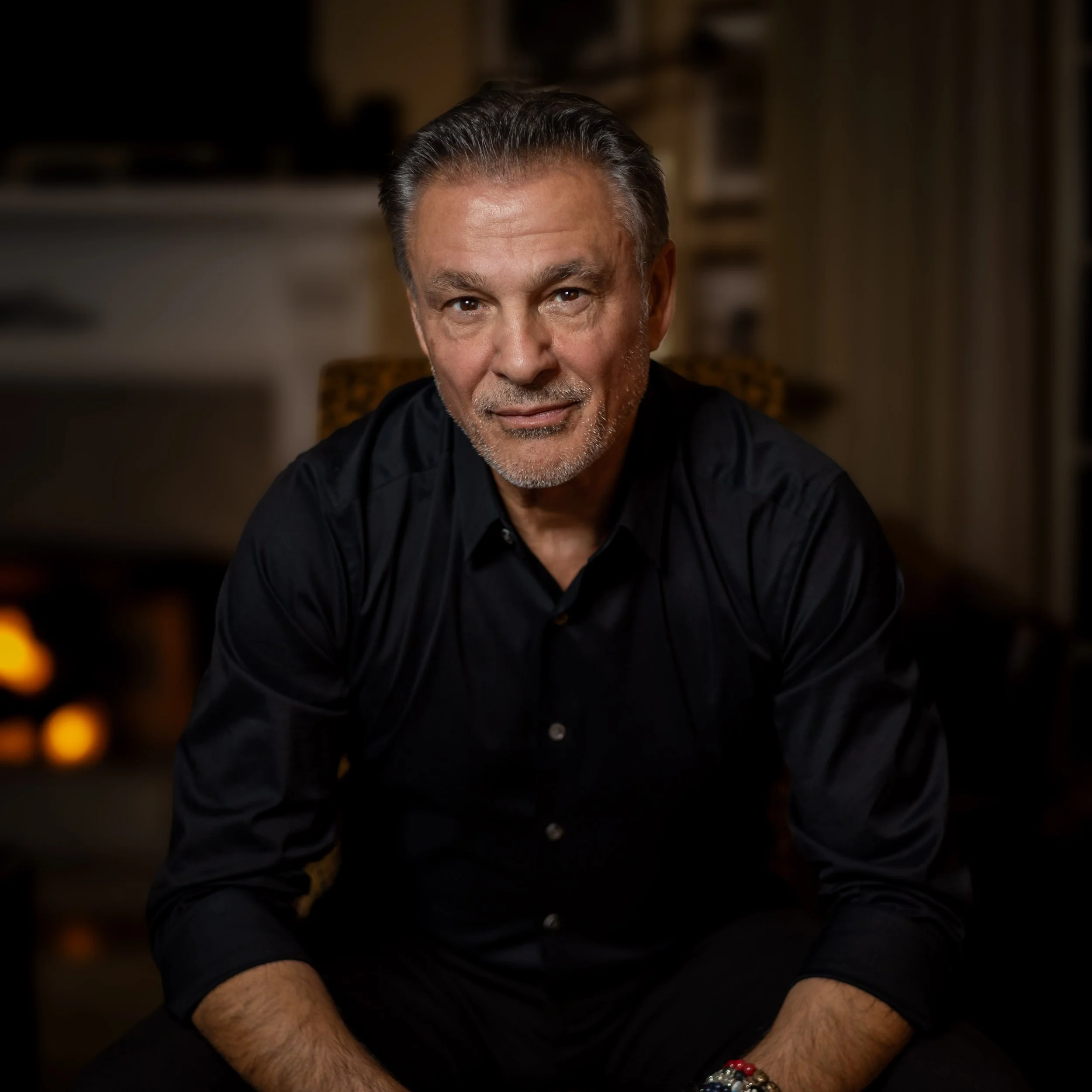 A middle-aged man with gray hair and a beard, wearing a black button-up shirt, sitting indoors in a warmly lit room, looking directly at the camera.