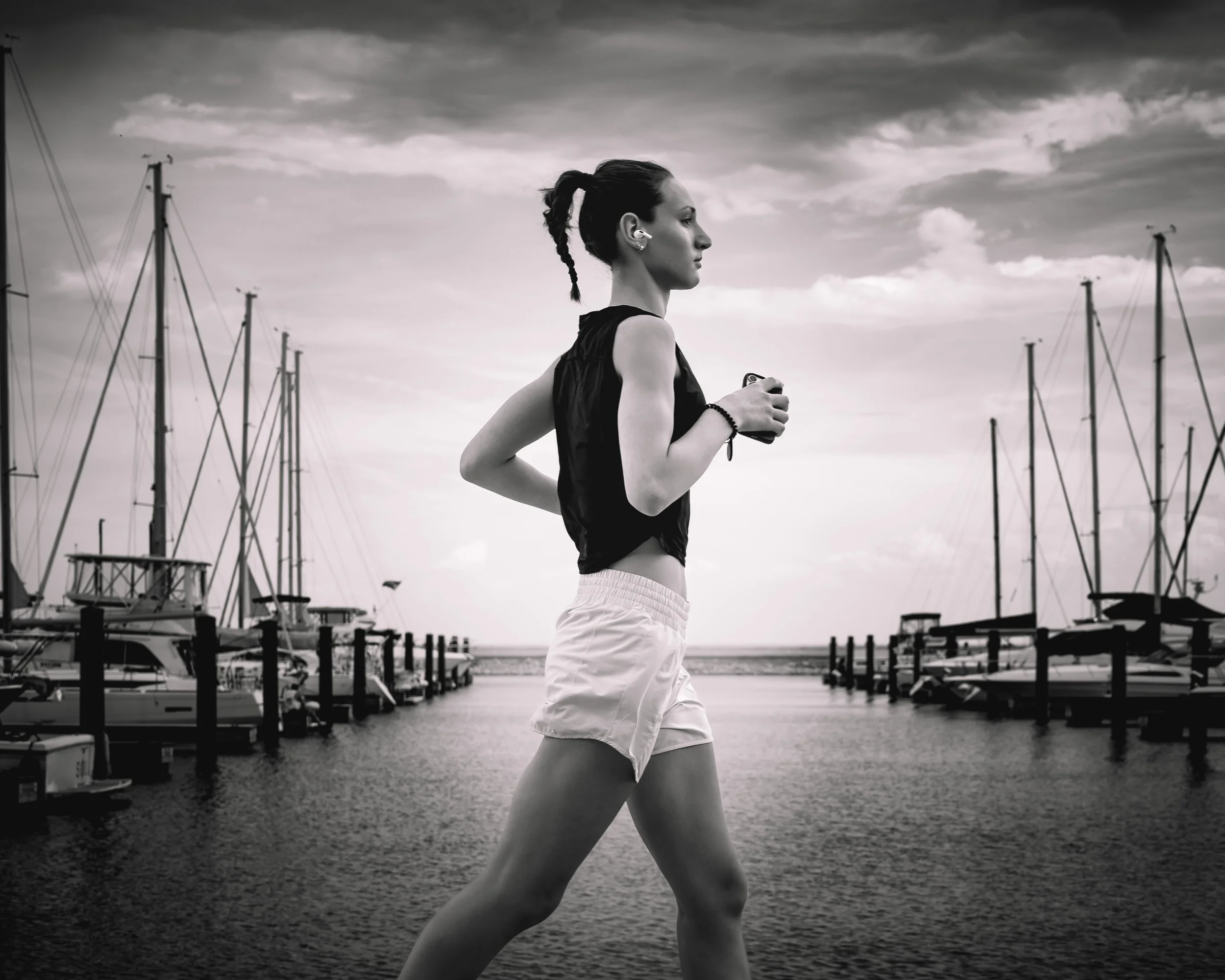 A woman jogging along a marina with sailboats docked on either side, in black-and-white.