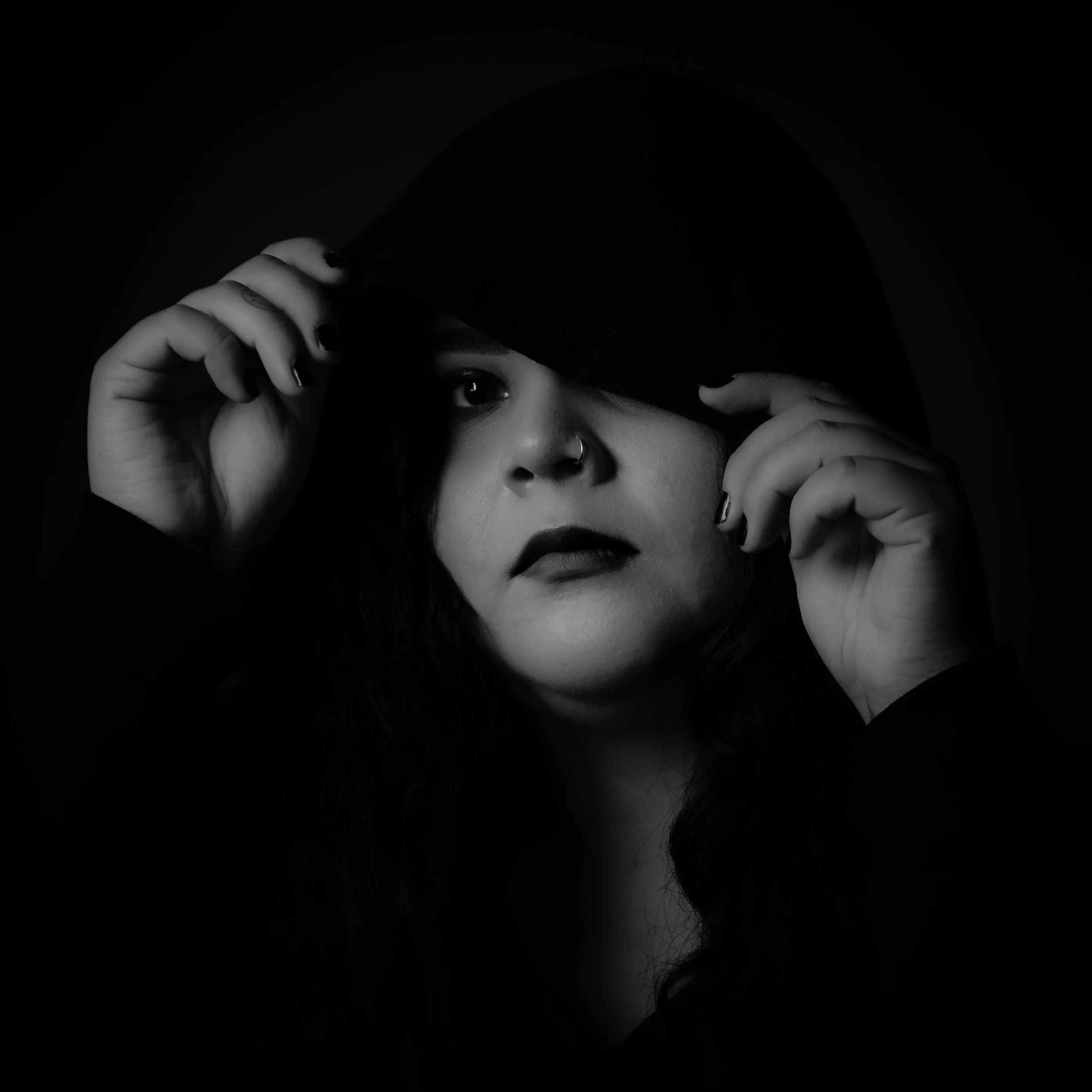 Black and white portrait of a woman with dark hair partially covering her face with a hood, holding the hood with both hands, looking directly at the camera.