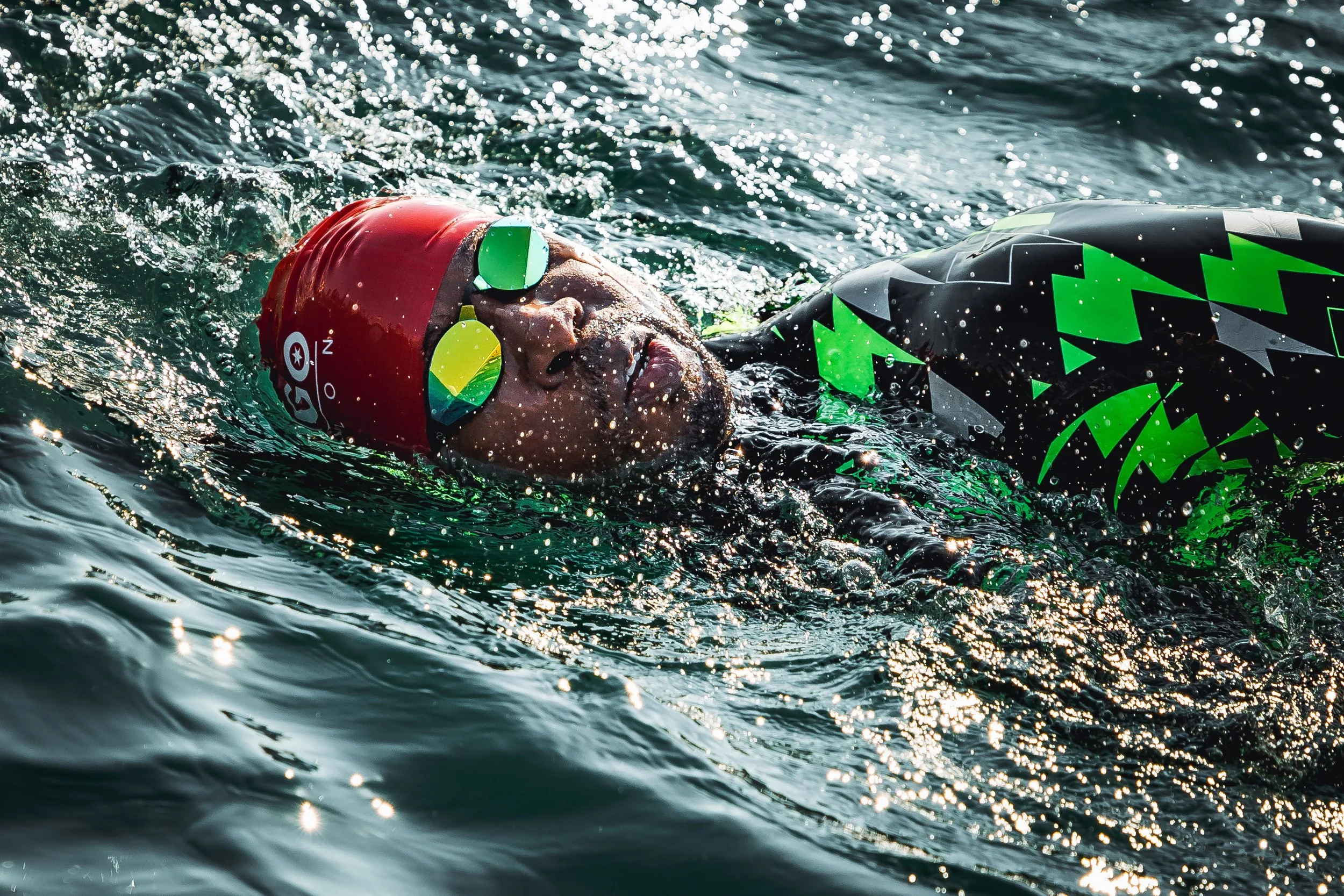 A man swimming in open water, wearing a red swim cap with a logo and mirrored sunglasses, in a black wetsuit with green and gray geometric patterns.