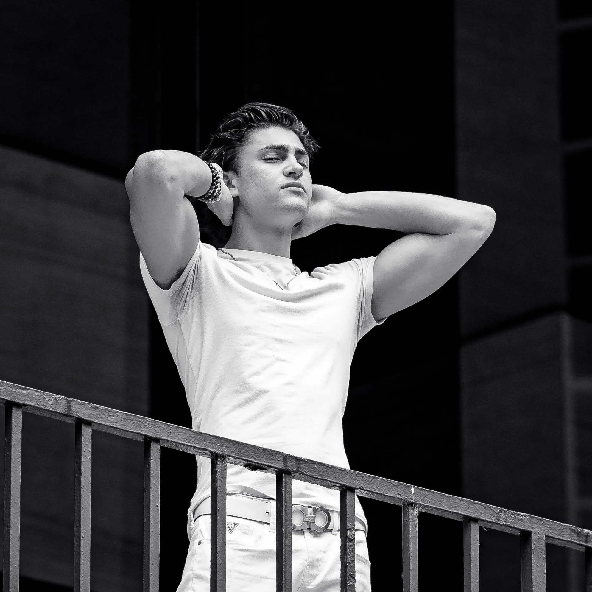 Black and white photo of a young man standing behind a railing, with his hands behind his head, wearing a white t-shirt and bracelets, looking confident.