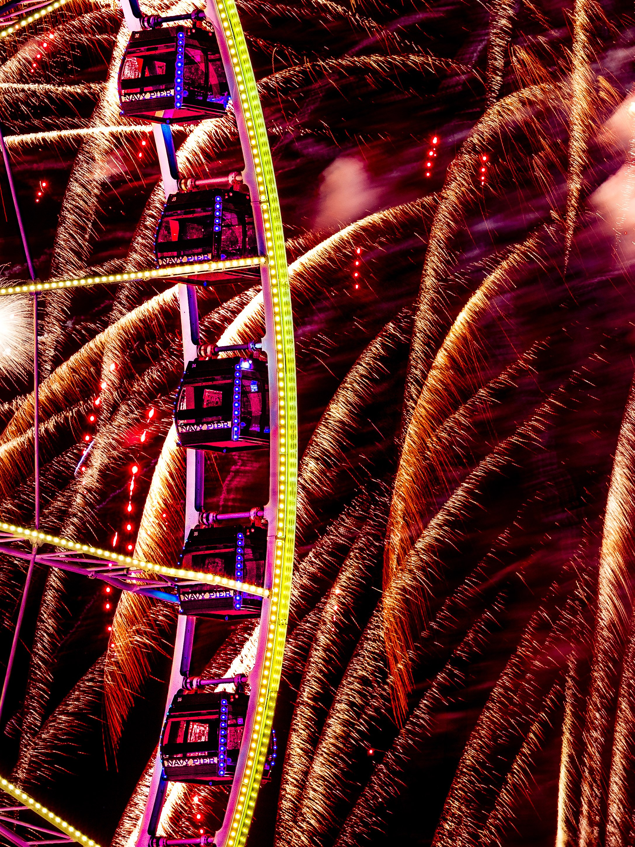 A brightly lit Ferris wheel at night, with fireworks exploding in the background.