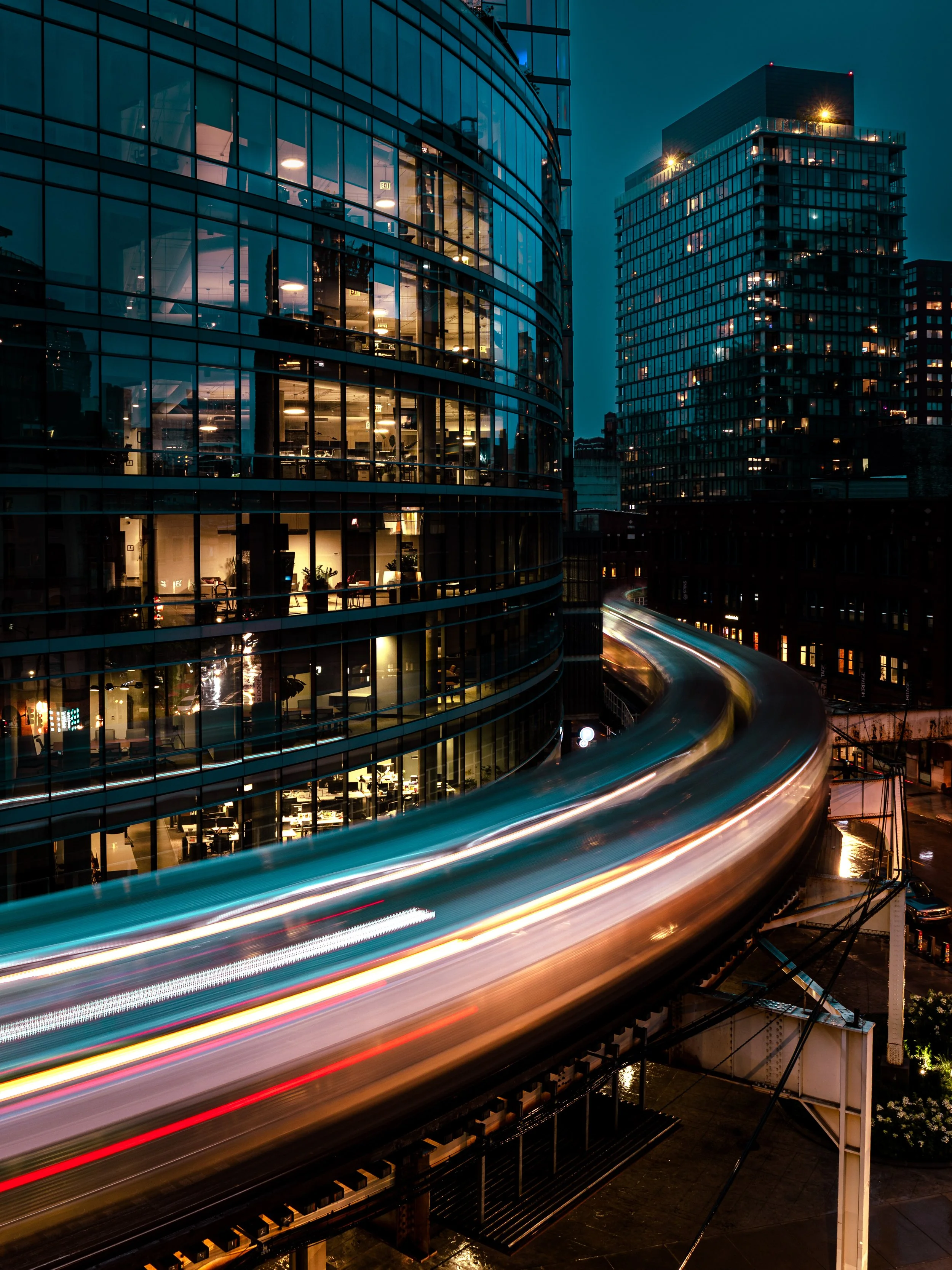 Nighttime city scene with glass office buildings and a curved CTA train passing in front, showcasing light trails from the moving train.