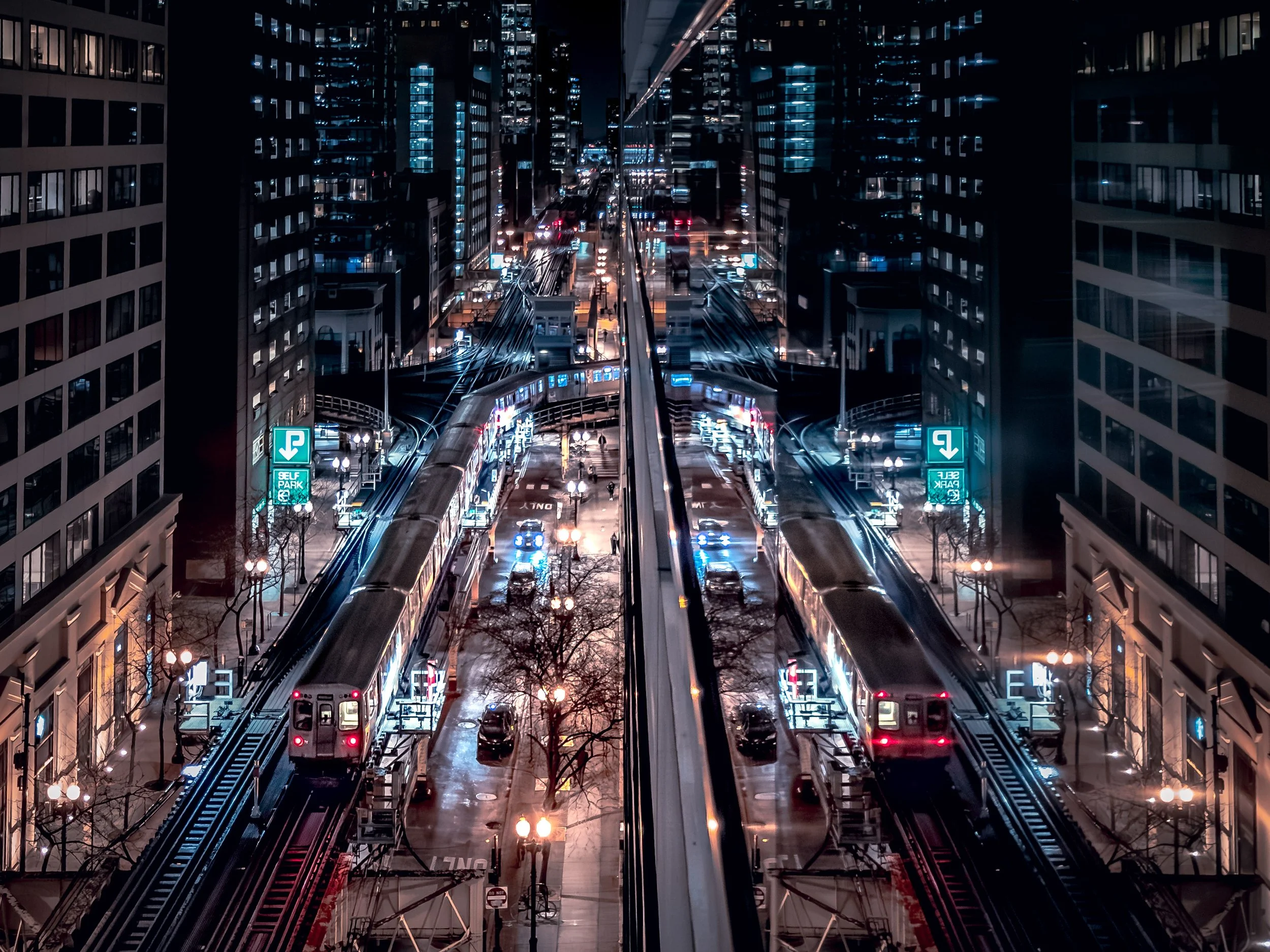 Night view of a city street with a train on elevated tracks, modern tall buildings, illuminated signs, and streetlights reflecting on wet pavement.