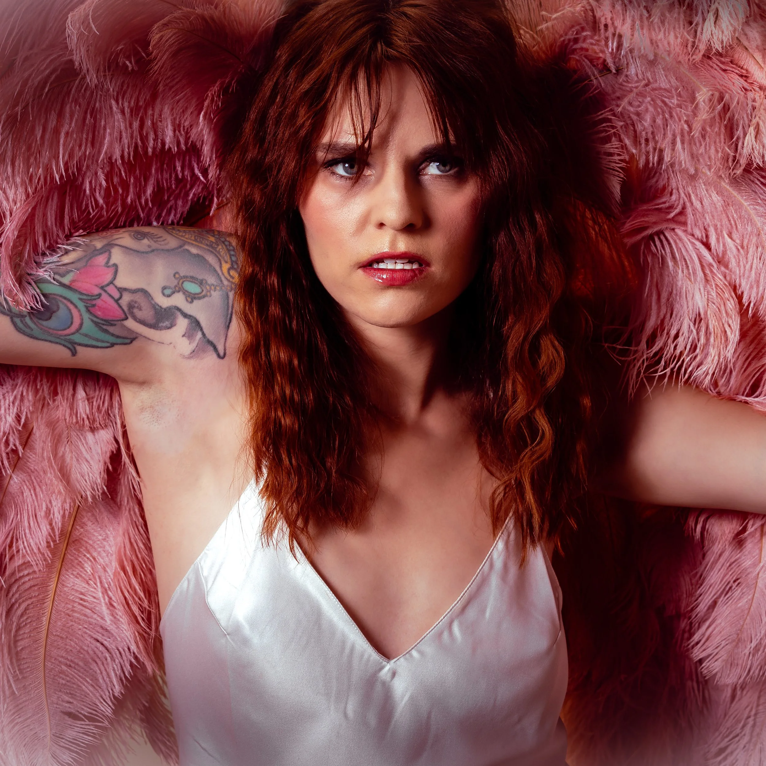 A woman with long, wavy red hair and tattoos on her arm, lying on pink feathers in a satin white top, with an intense expression.