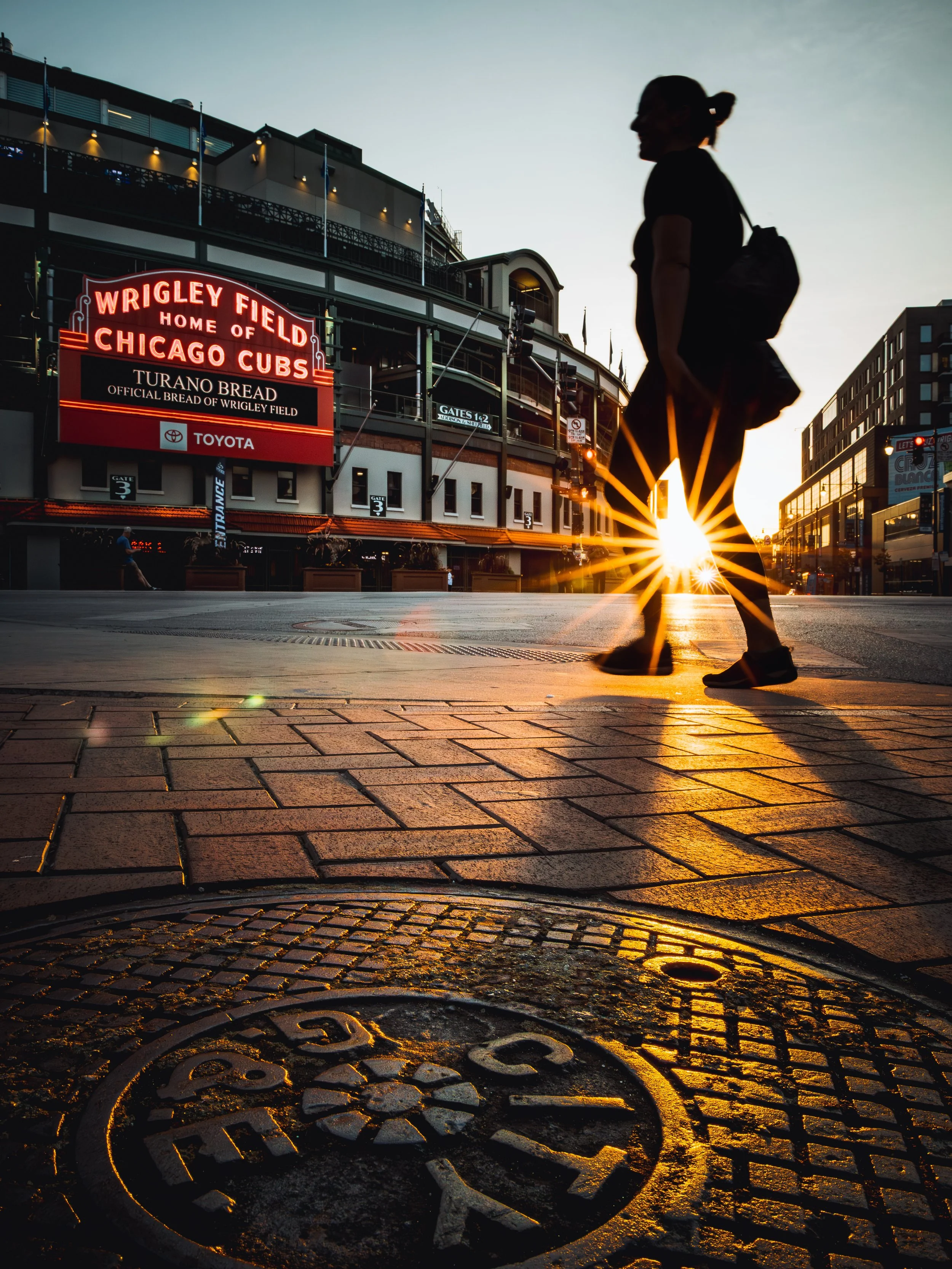 A person walking in front of Wrigley Field in Chicago at sunrise, with a woman silhouette, street scene, and the sun shining through her legs.