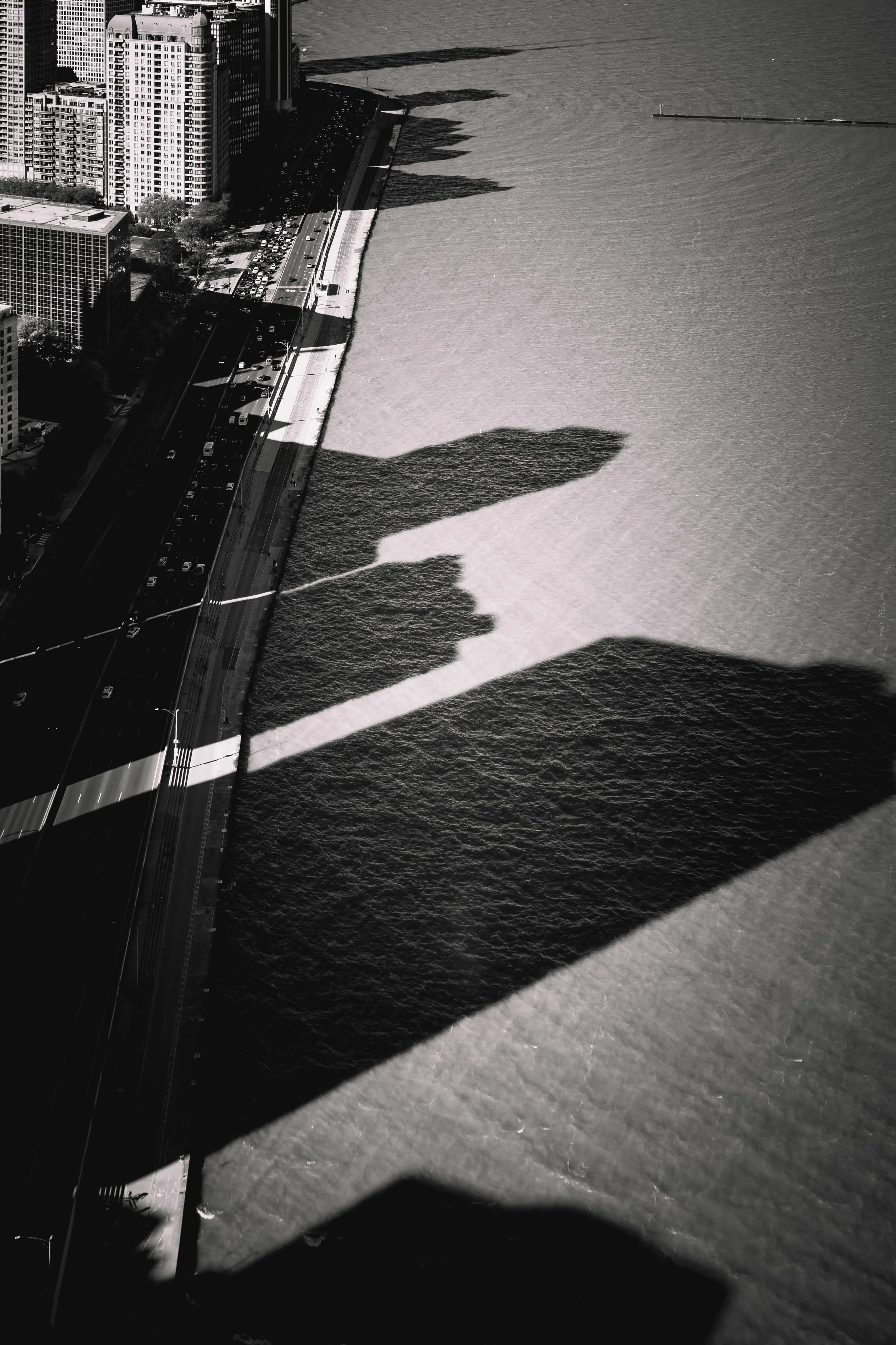 Black and white aerial photo of a city shoreline showing tall buildings, a busy road with cars, and the water's edge casting a shadow from a bridge or structure over the water.