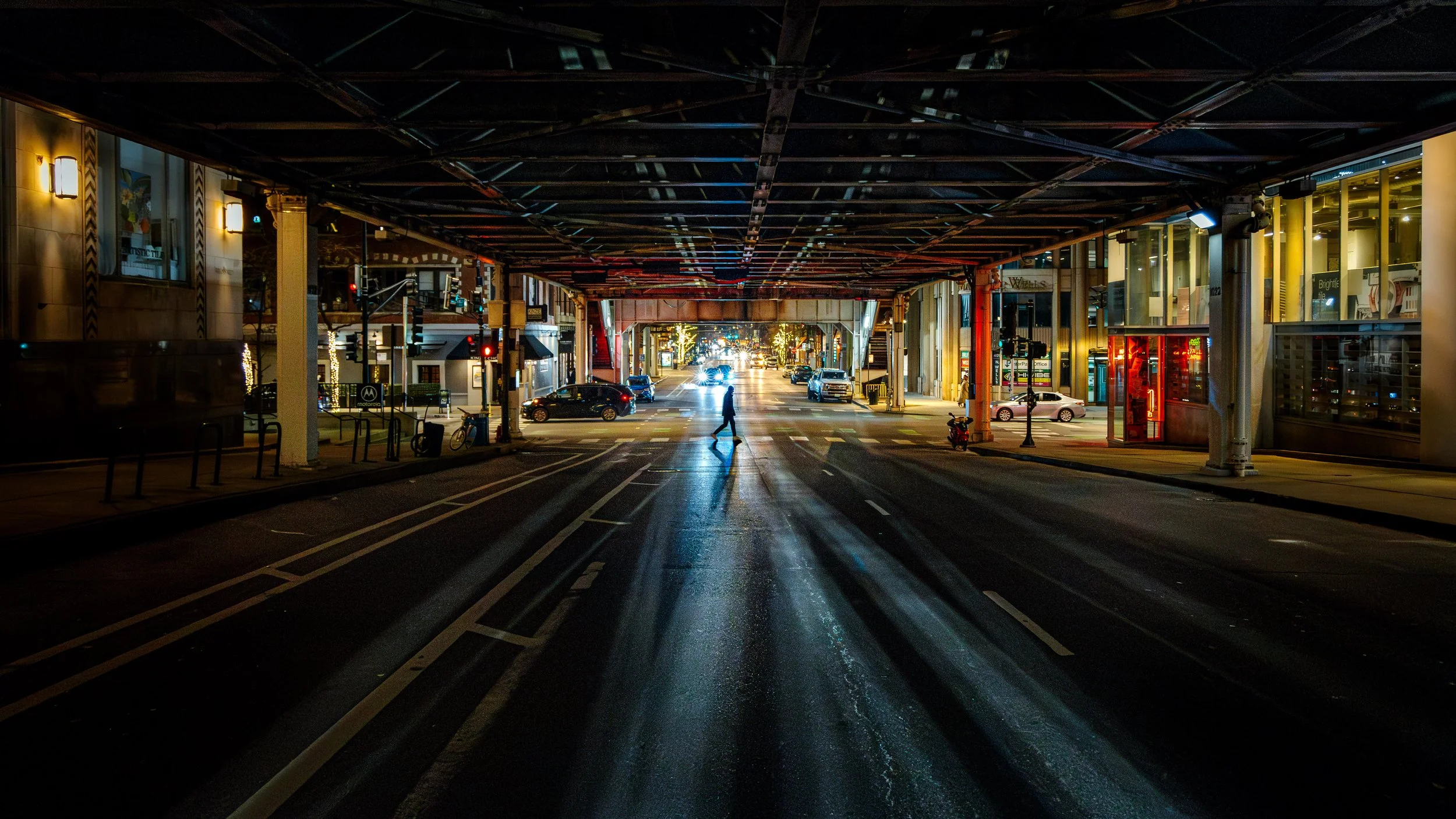 A city street at night viewed from under an overpass. A person crosses the street, with cars and bicycles parked along the sides. Buildings with lit windows line both sides, and streetlights illuminate the scene with a festive atmosphere.