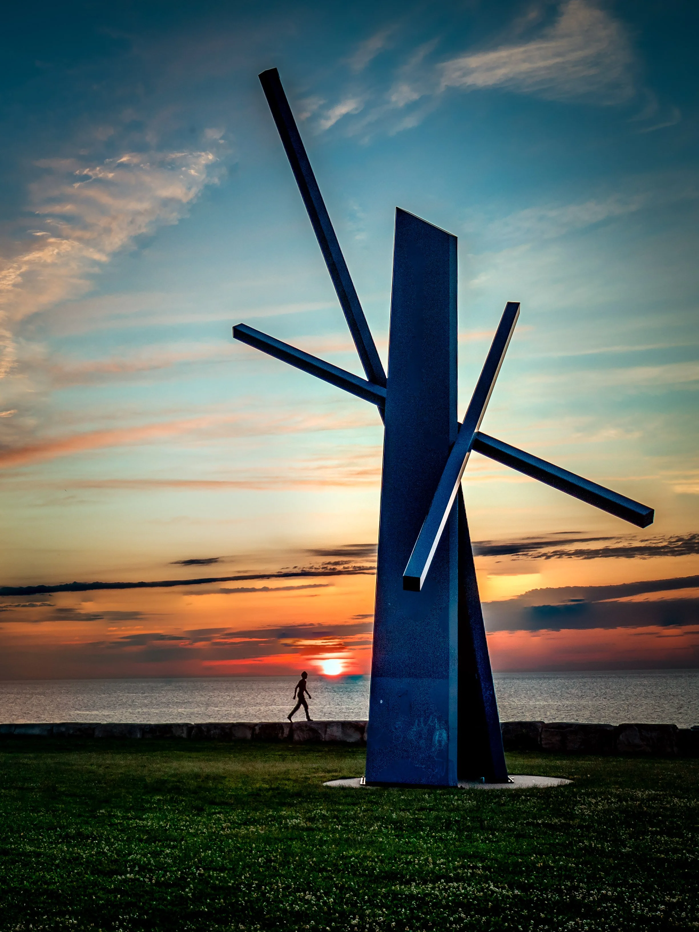 The Chicago Chevron sculpture outdoors near the water at sunrise with a person walking along the shoreline and colorful sky