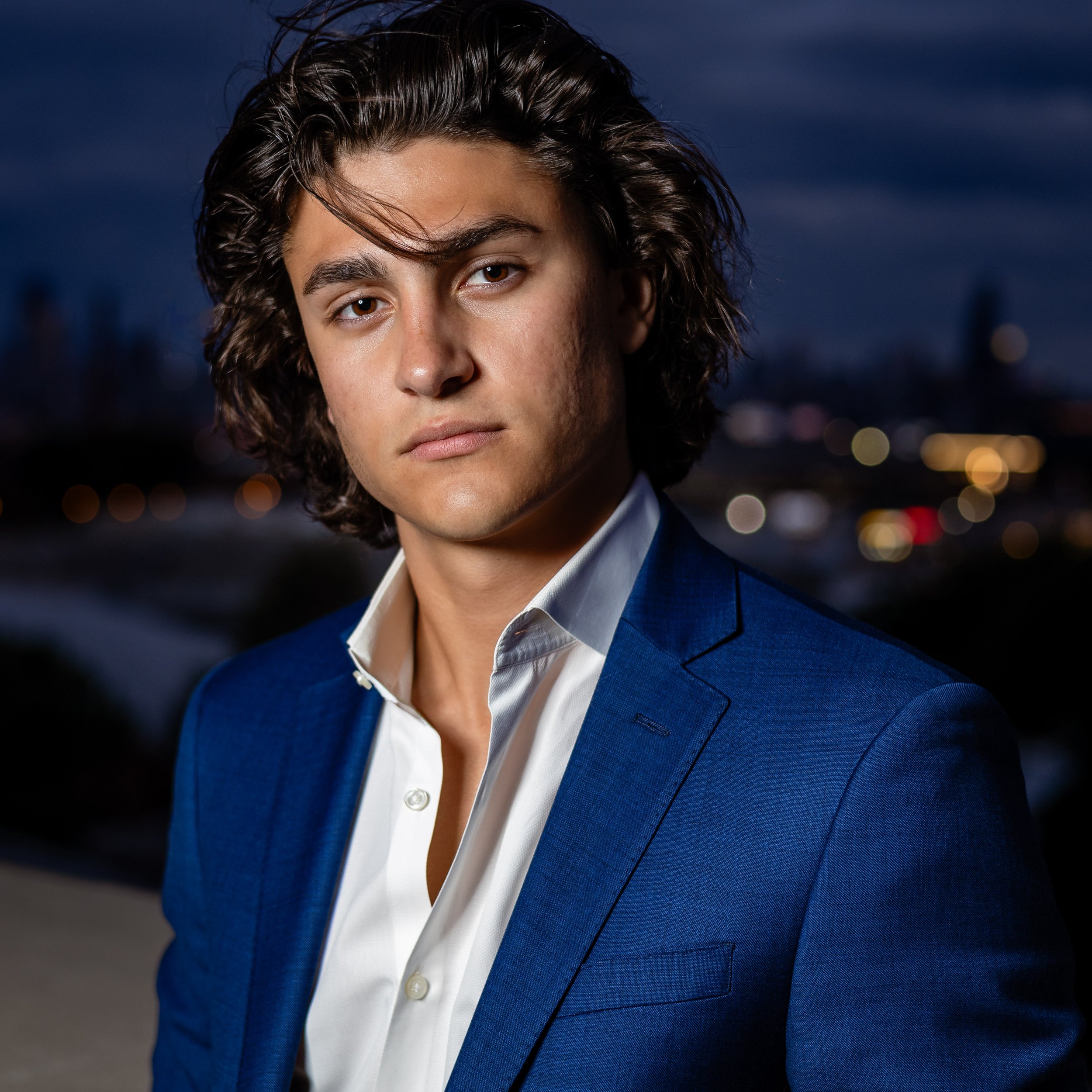 Young man with wavy dark brown hair wearing a blue suit with a white dress shirt, standing outdoors at night with city lights in the background.