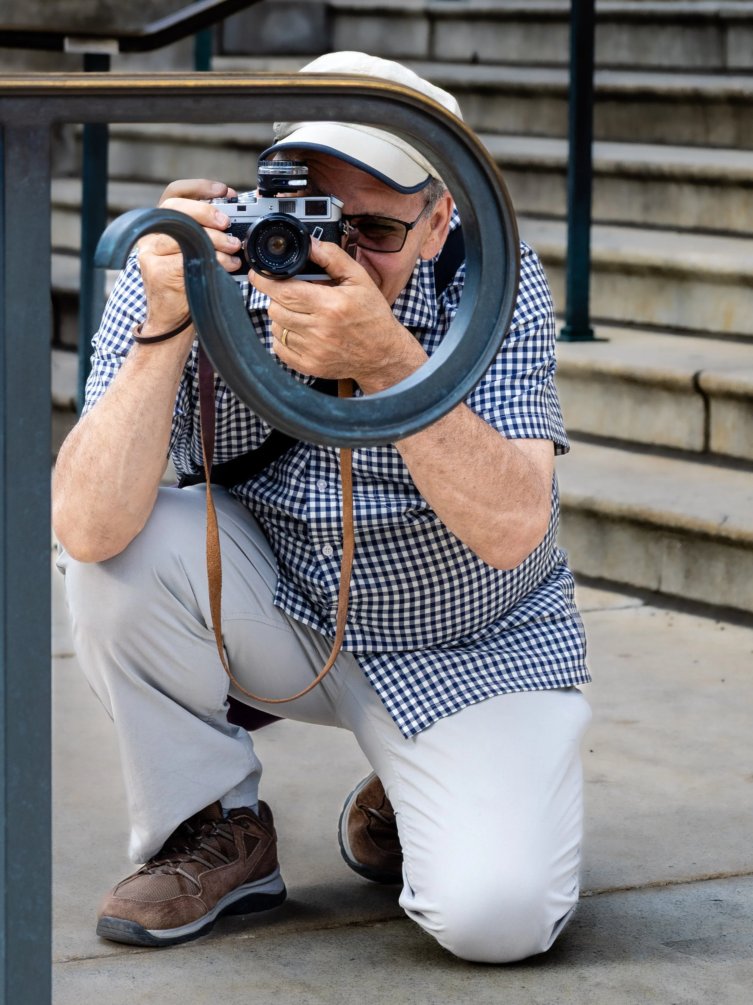 A man crouching on the ground taking a photo through a circular railing with a camera. He is wearing a checkered shirt, beige pants, brown shoes, glasses, and a beige hat.