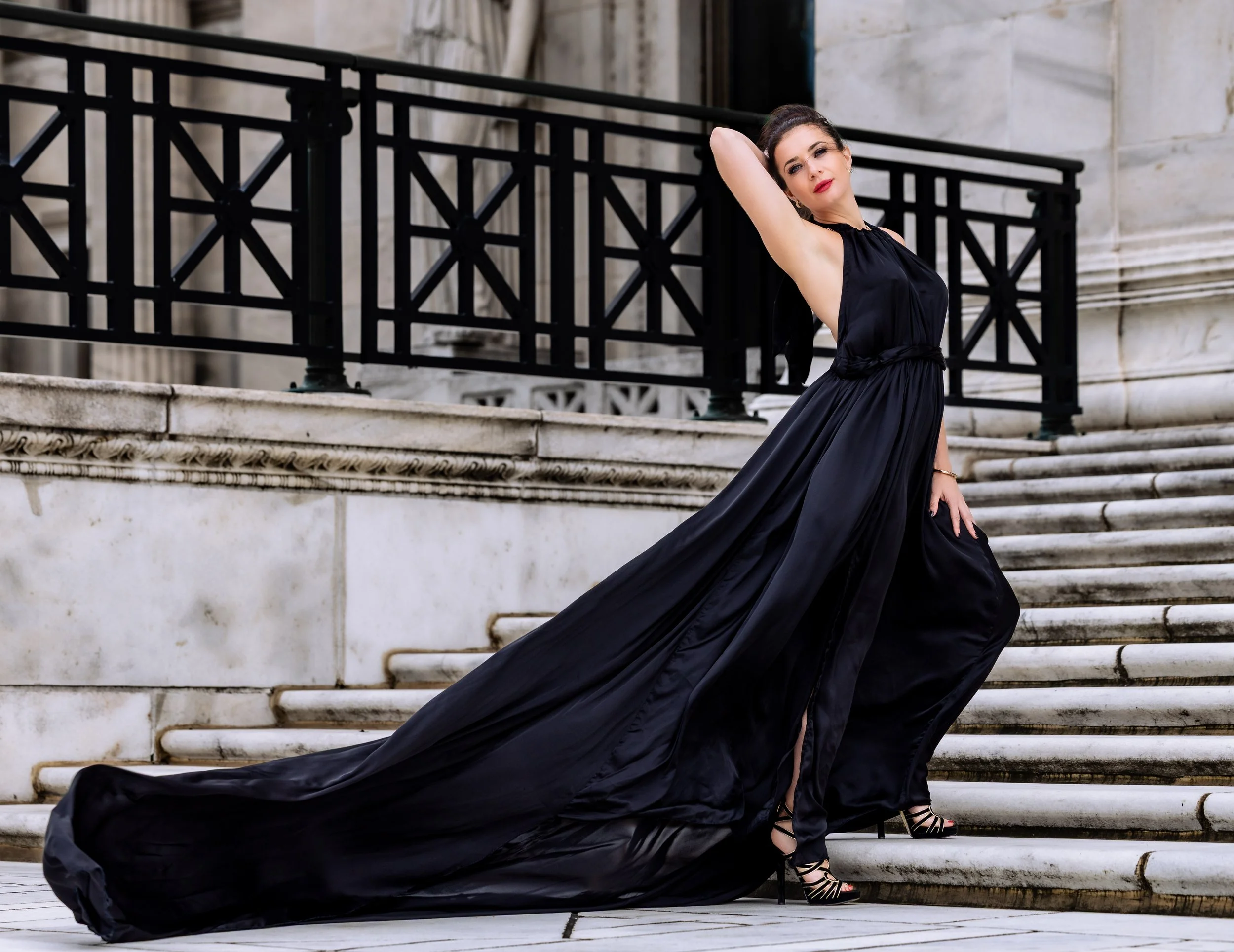 A woman in a long black gown with a train, posed on marble stairs in front of a building with stone walls and black metal railing.