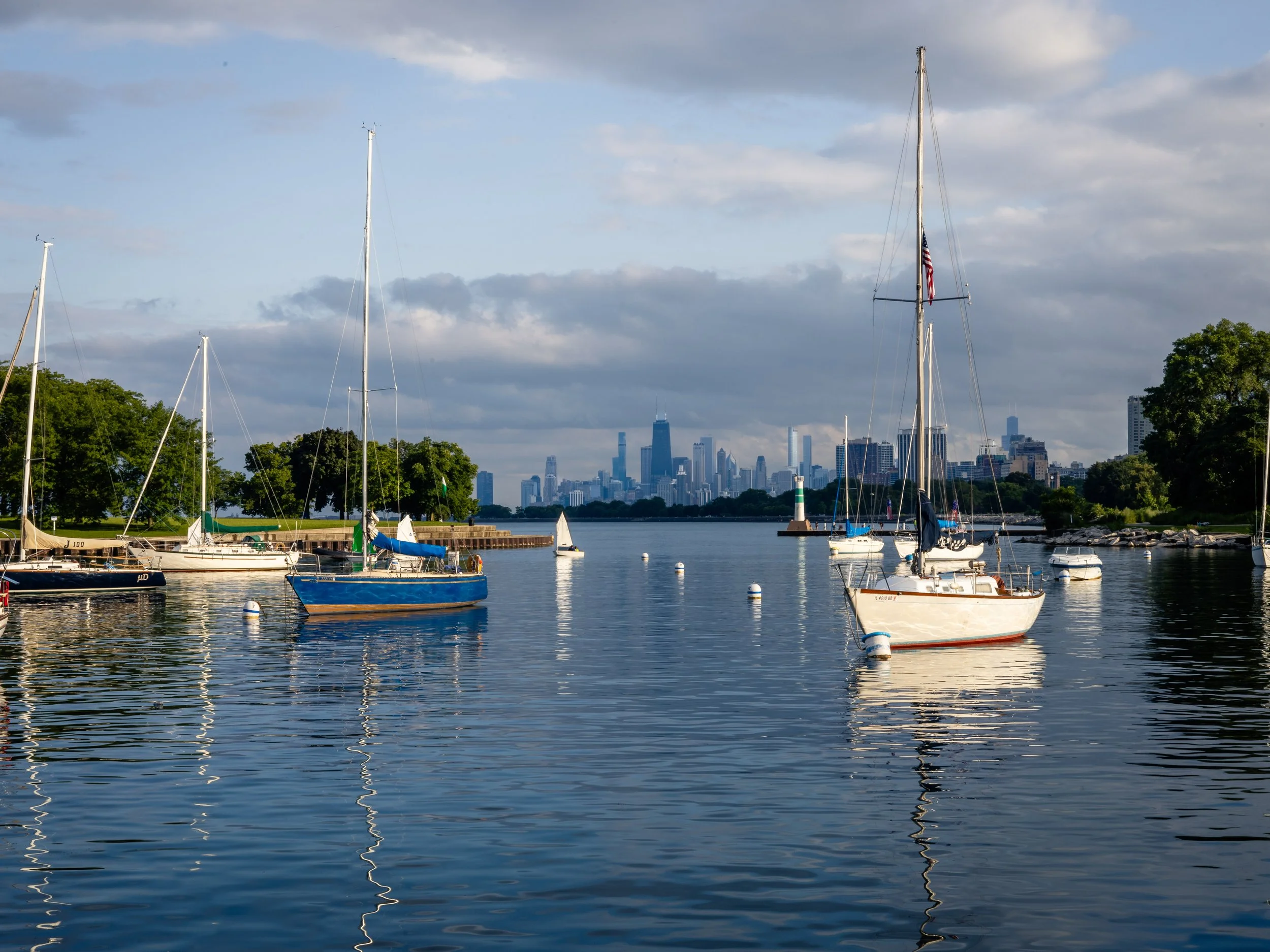 Boats anchored in a peaceful harbor with a city skyline in the distance on a partly cloudy day.