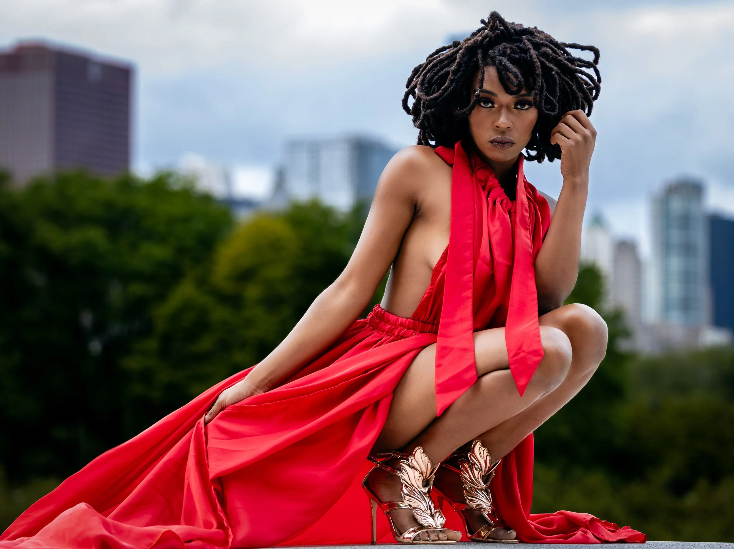 A woman with dreadlocks posing outdoors in a red outfit and golden high-heeled shoes, with a cityscape and green trees in the background.