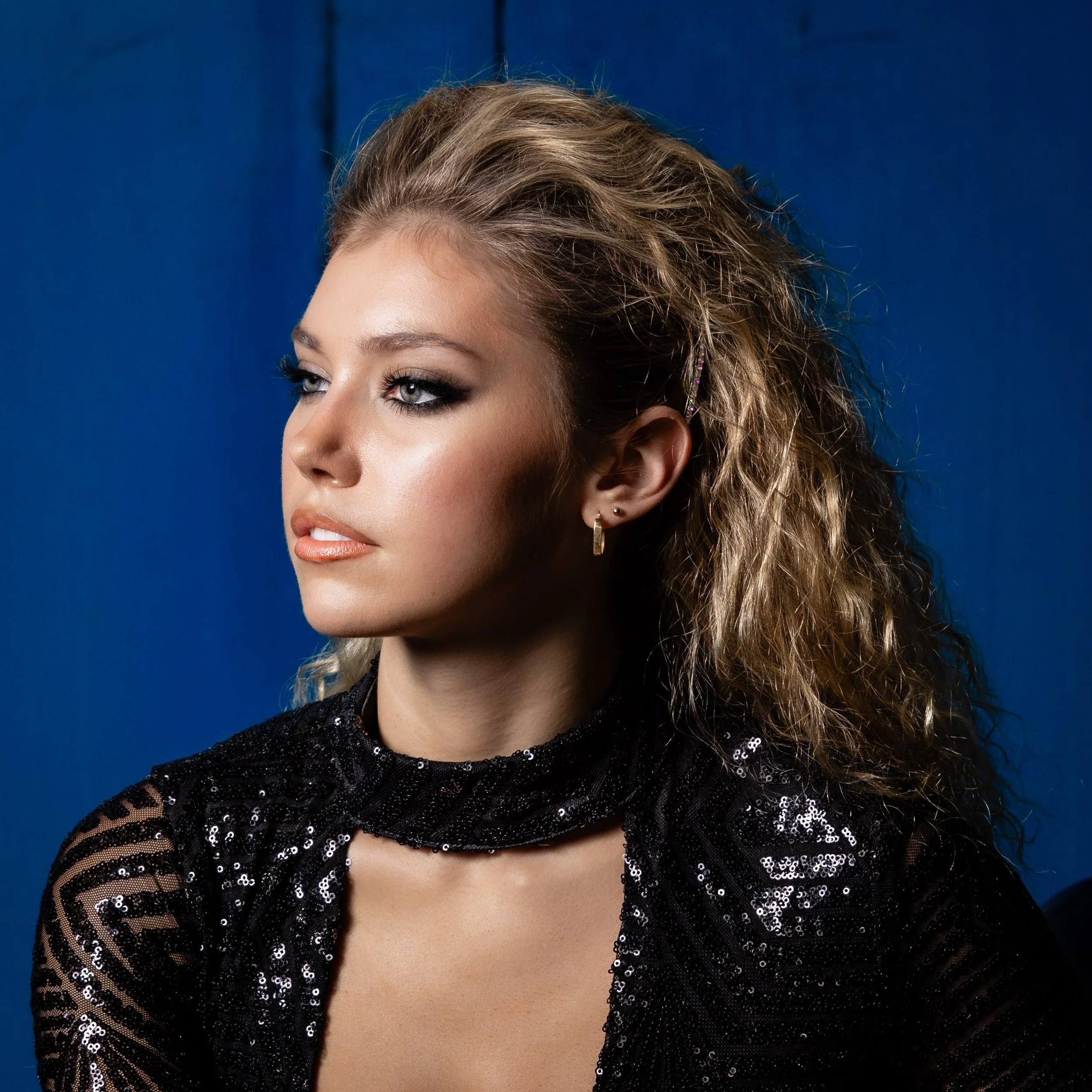 A young woman with curly blonde hair, wearing earrings, a black sequin top, and light makeup, looking to the side against a blue background.