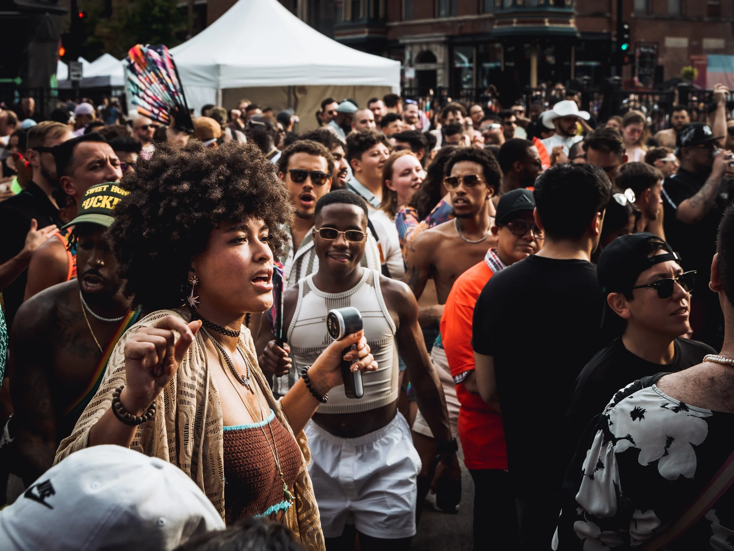 Crowd of diverse people at outdoor event with tents and a building in background, many wearing sunglasses and casual attire, some holding fans or microphones.