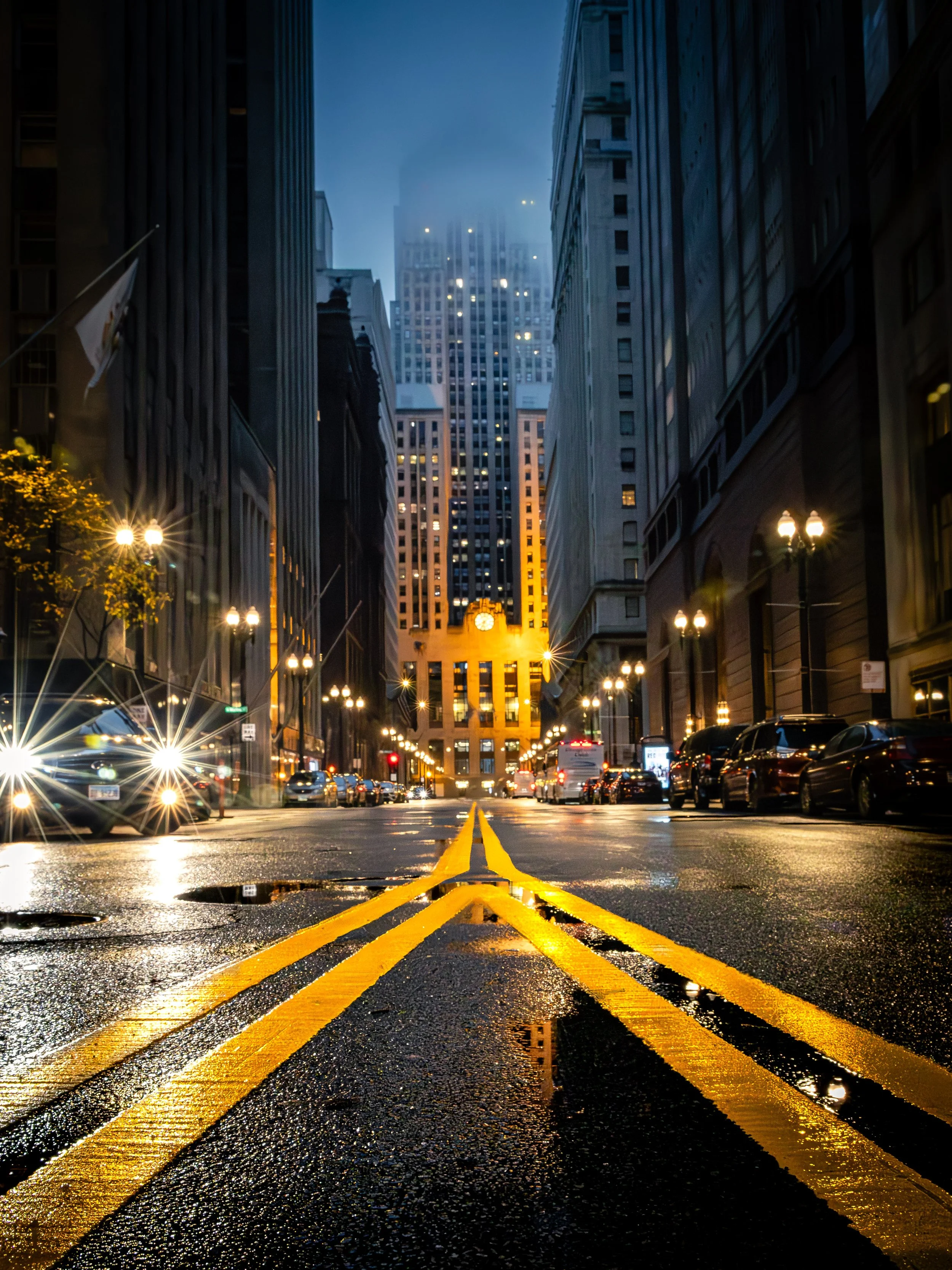 City street at night with wet pavement, yellow road lines, tall buildings on both sides, and a lit clock tower at the end of the street.