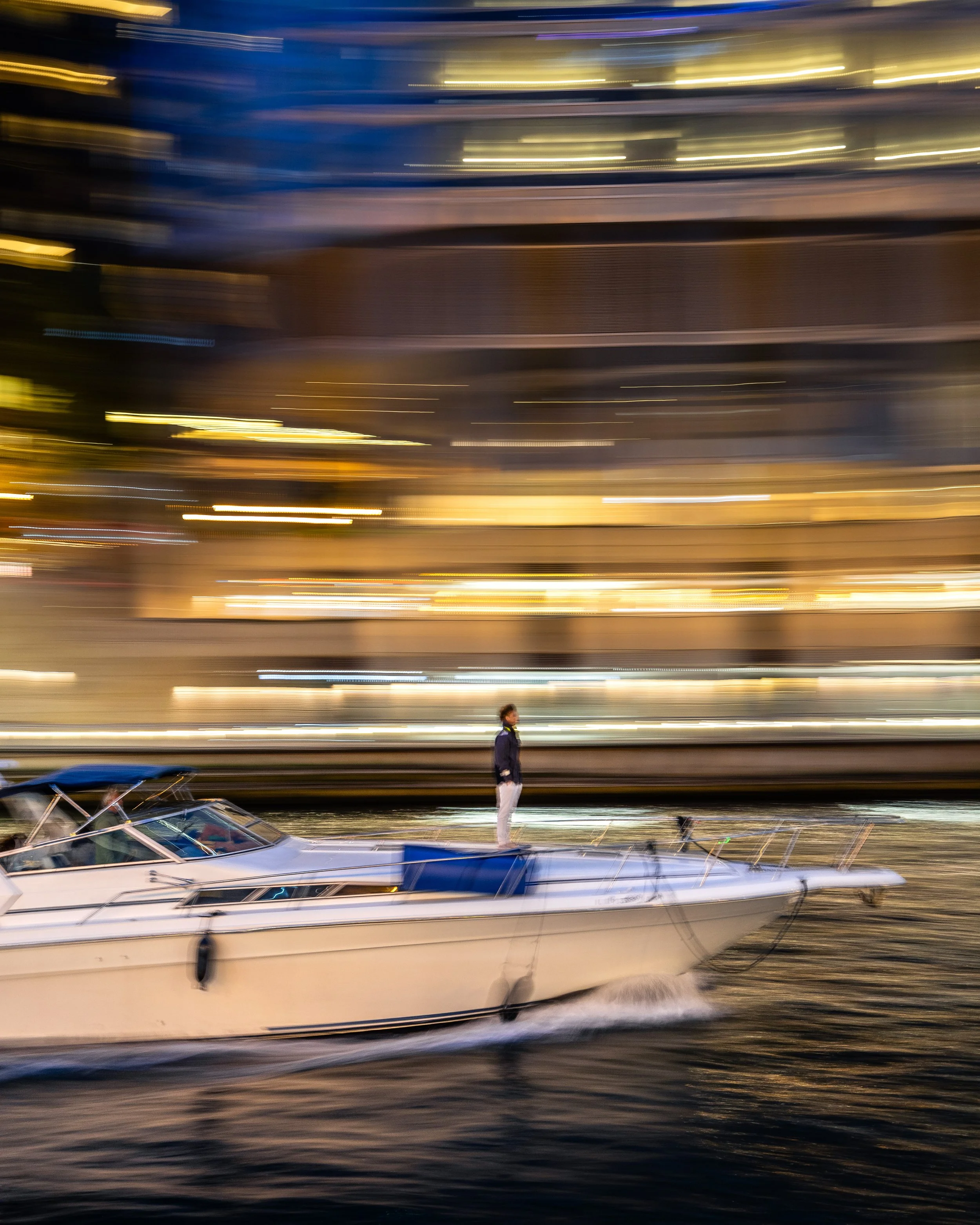 A person standing on a boat on water at night with blurred city lights in the background.