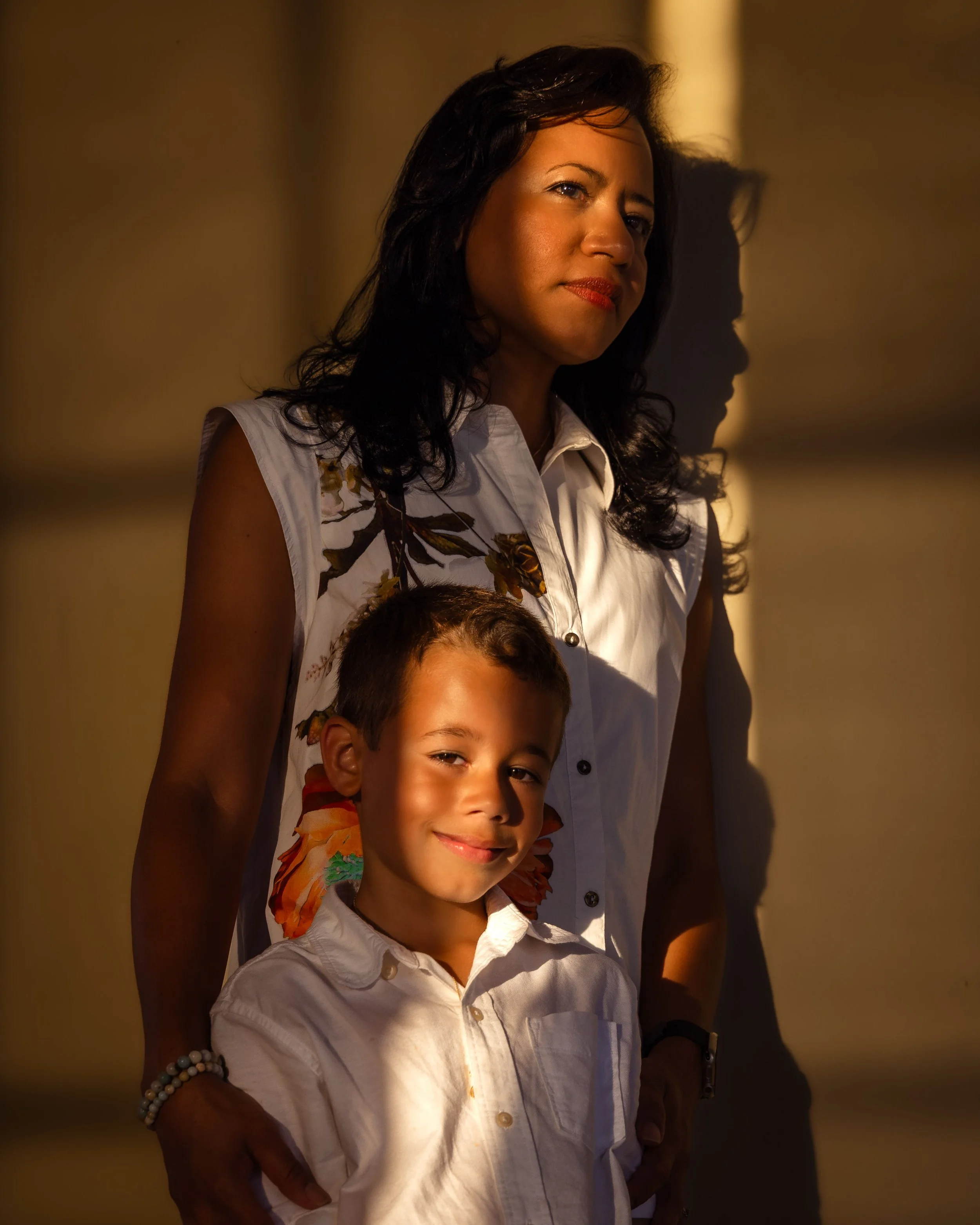 A woman and a boy standing against a wall illuminated by warm sunlight.