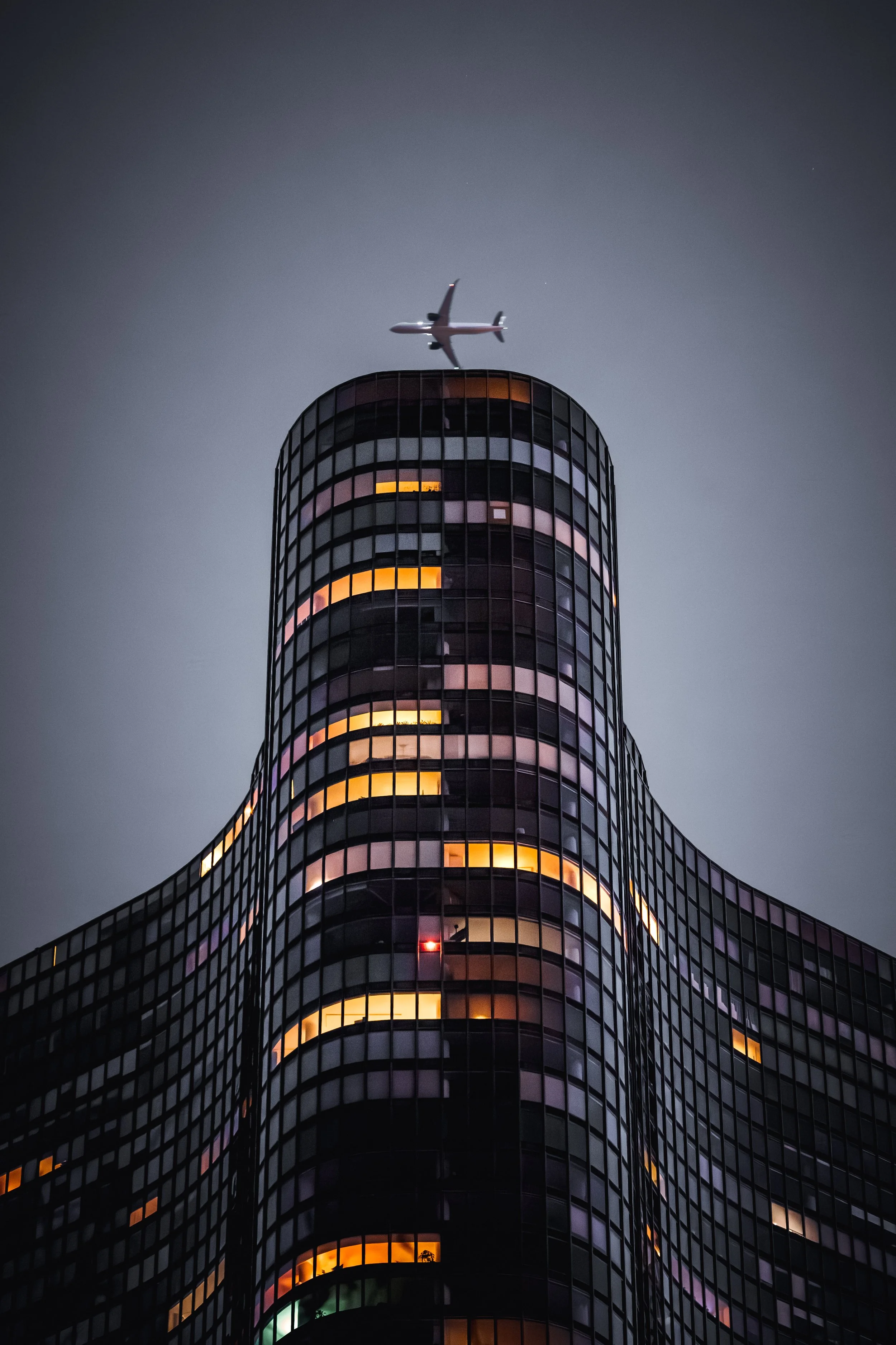 Nighttime shot of a tall, modern glass skyscraper with illuminated windows and an airplane flying overhead.