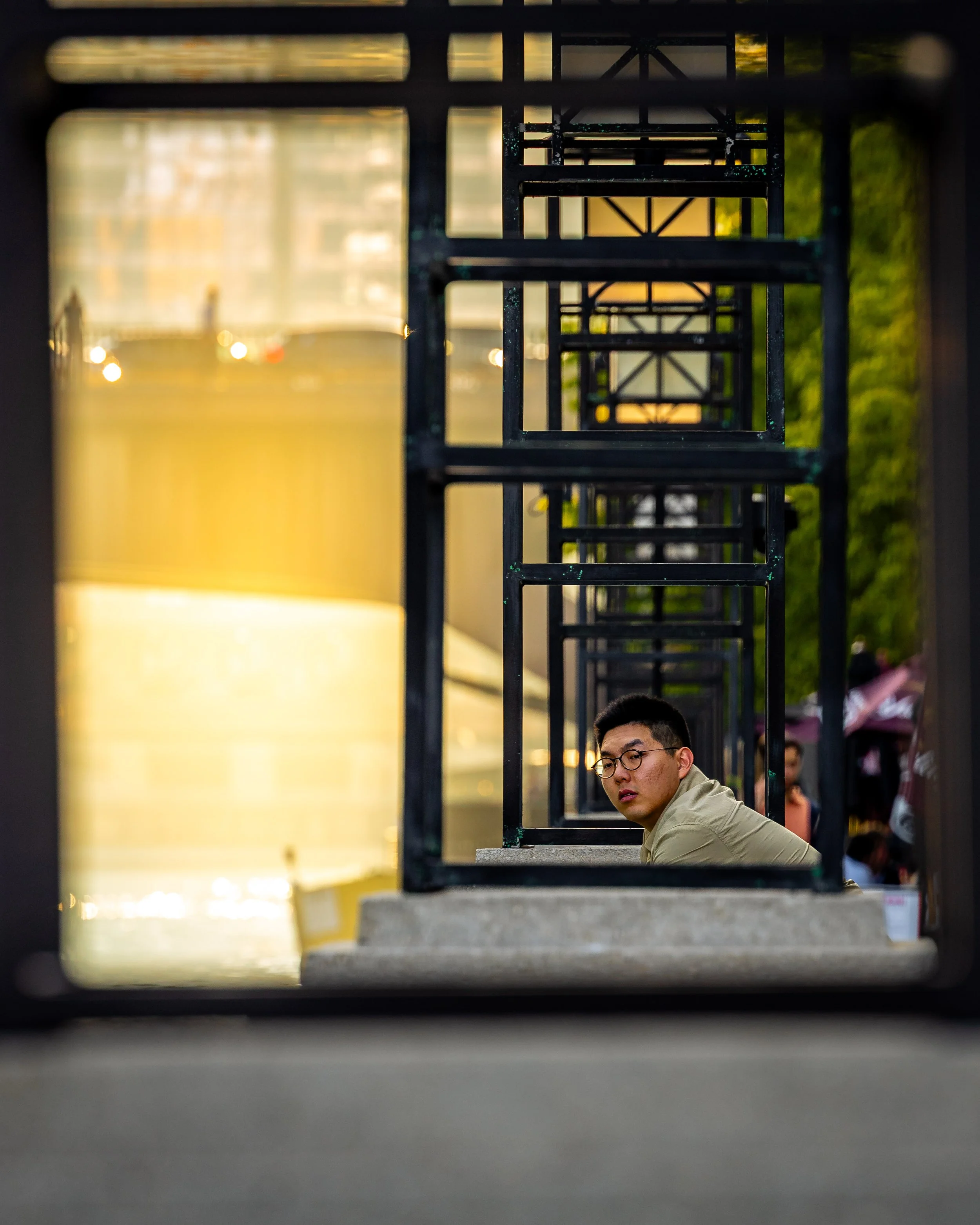 A young man wearing glasses and a beige jacket sitting on a concrete step, looking through a series of black metal rectangular frames that create a tunnel effect outside, with a blurred background of trees and people.