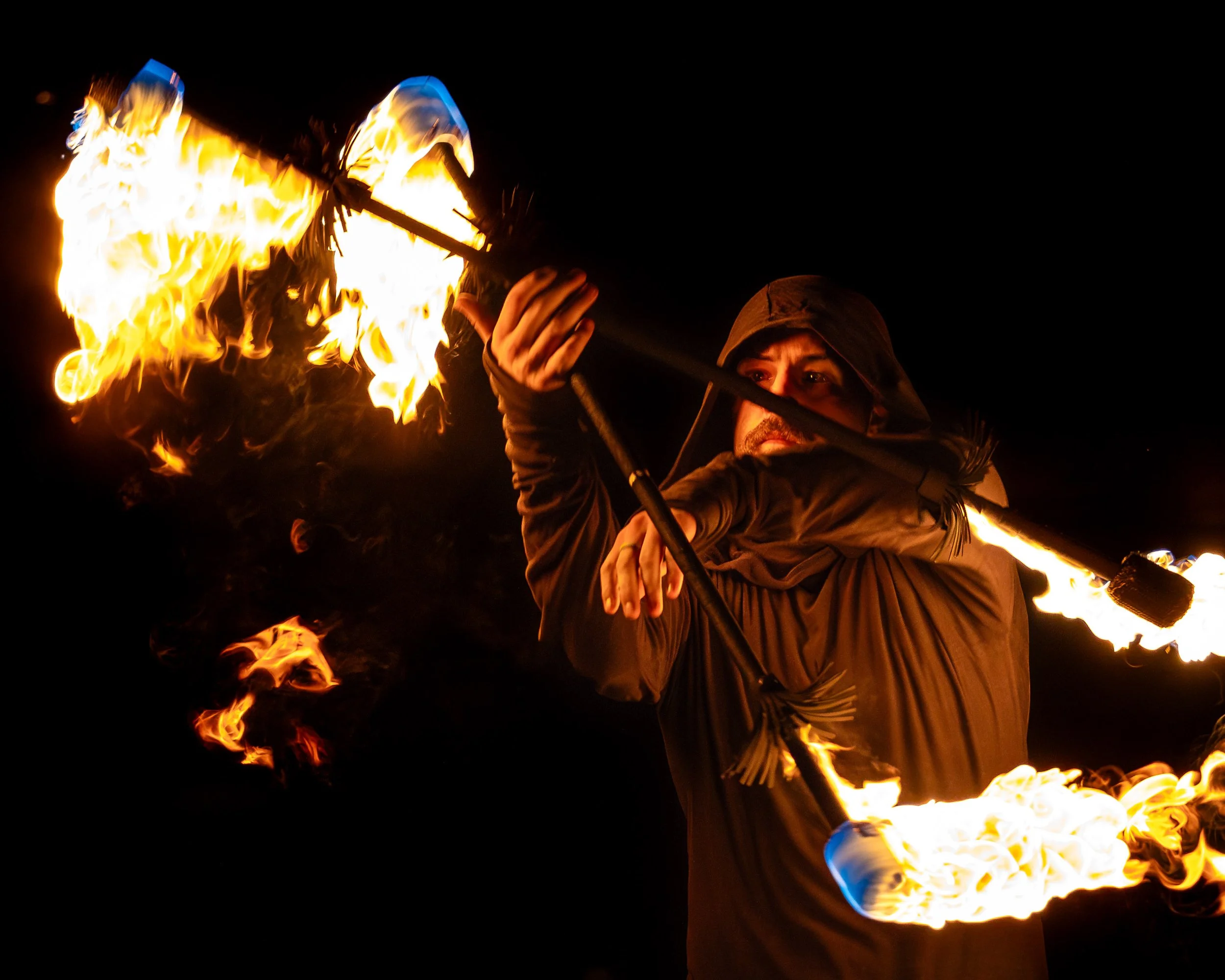 A person in a hooded jacket performing a fire poi act at night, swinging fire torches in a circular motion.