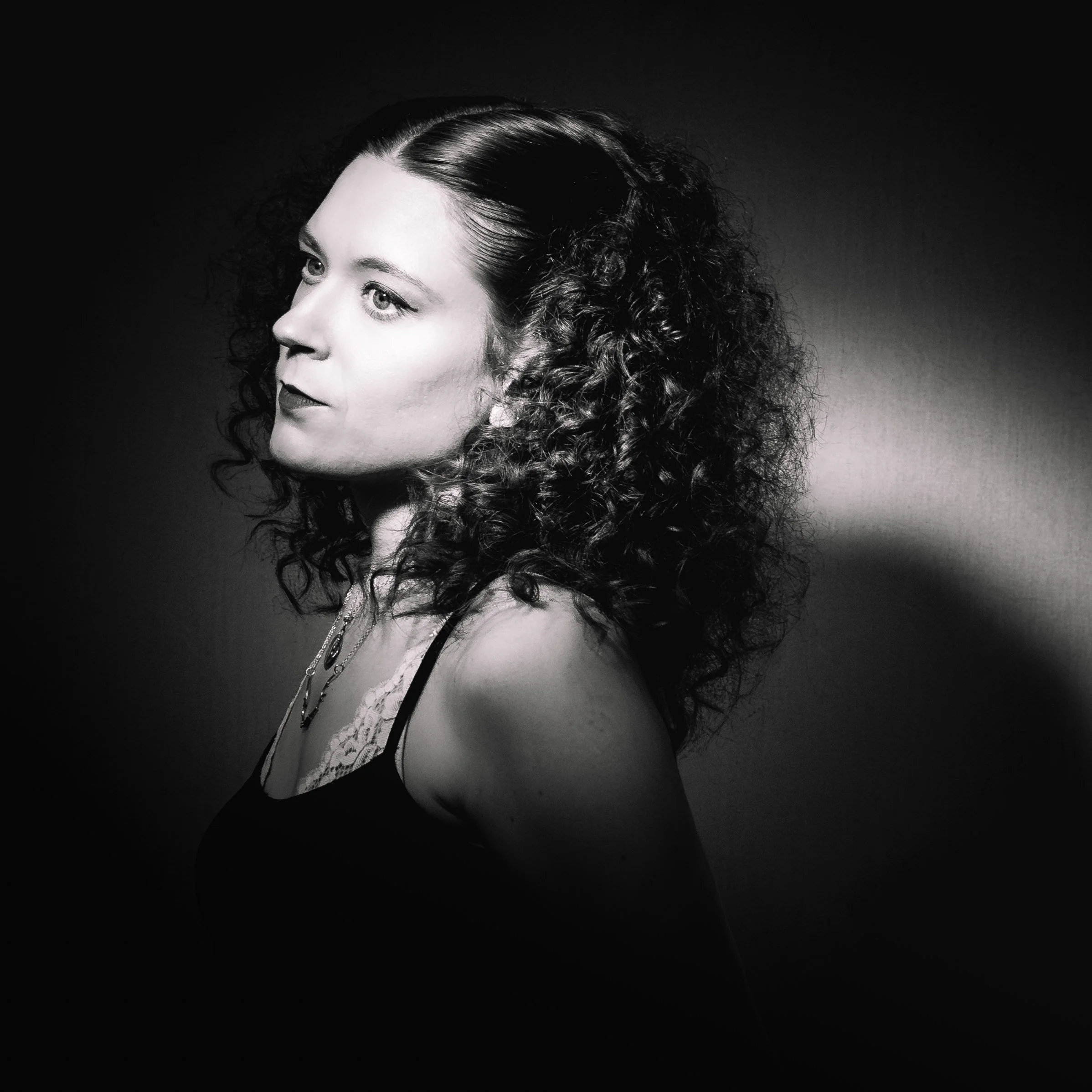Black and white portrait of a woman with curly hair, wearing a sleeveless top and a necklace, looking to the side.