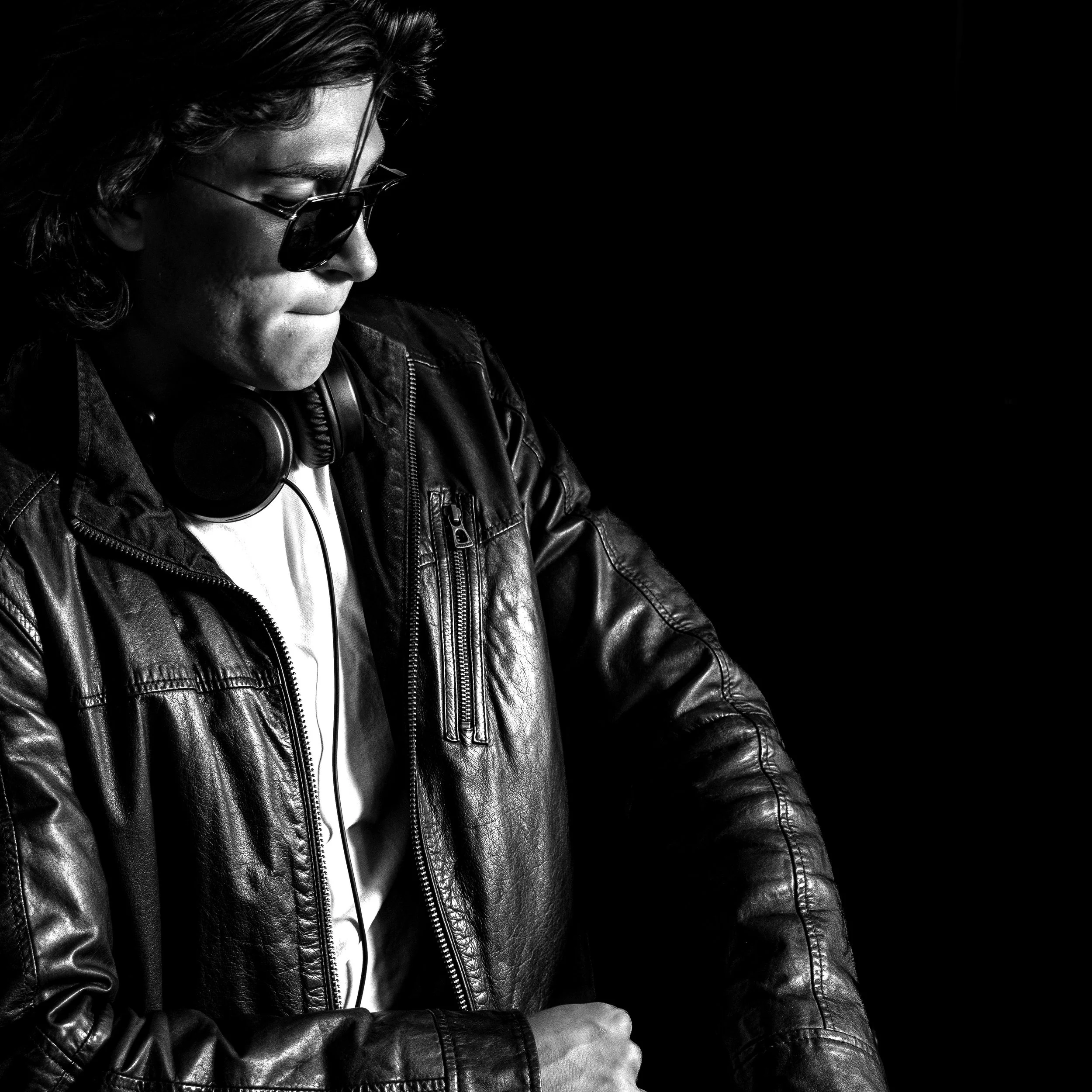 A man wearing sunglasses, a leather jacket, and headphones, looking down with a serious expression, against a black background.