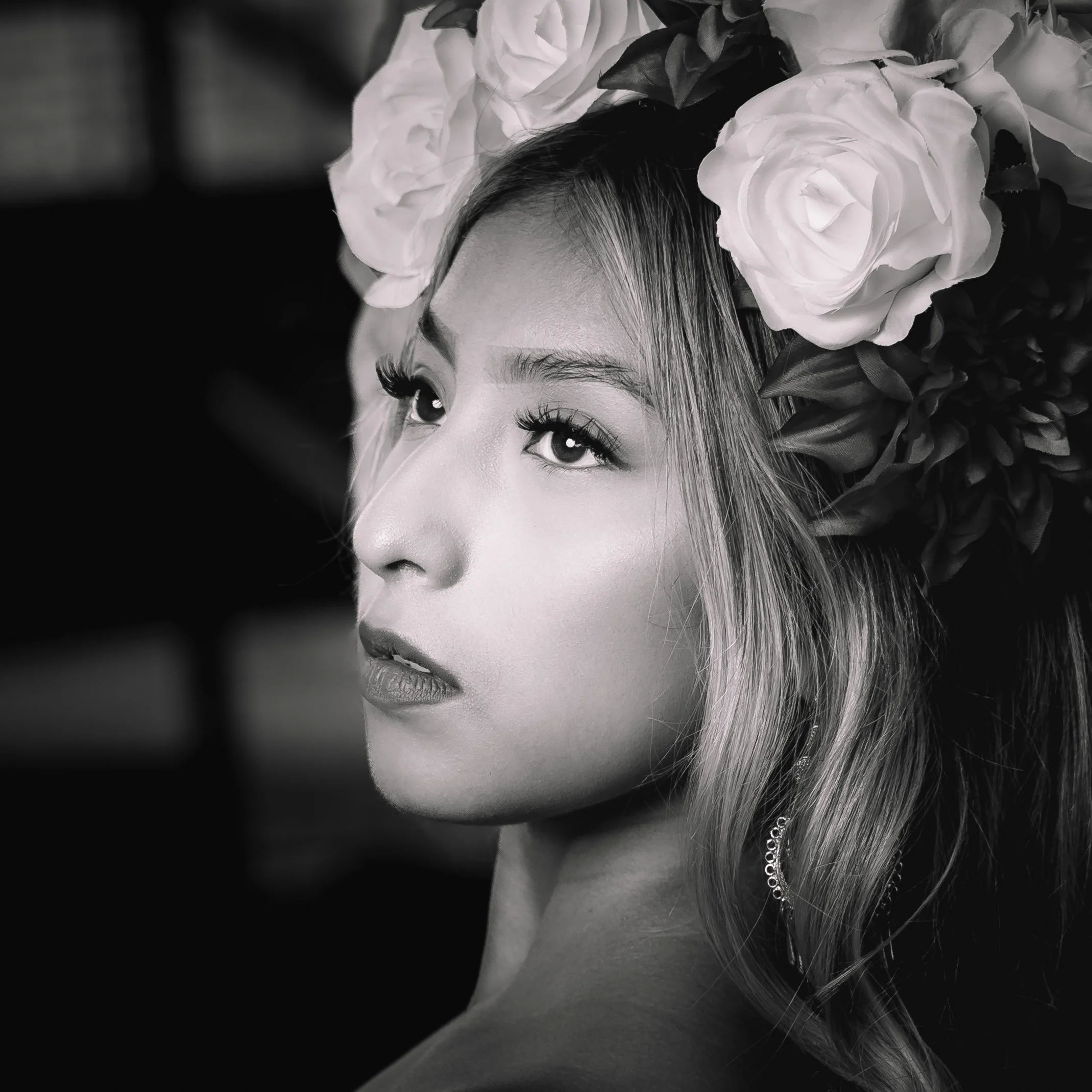 A black and white portrait of a woman with a floral headdress, featuring large roses and leaves. She has long eyelashes, smooth skin, and her gaze is directed slightly upward.