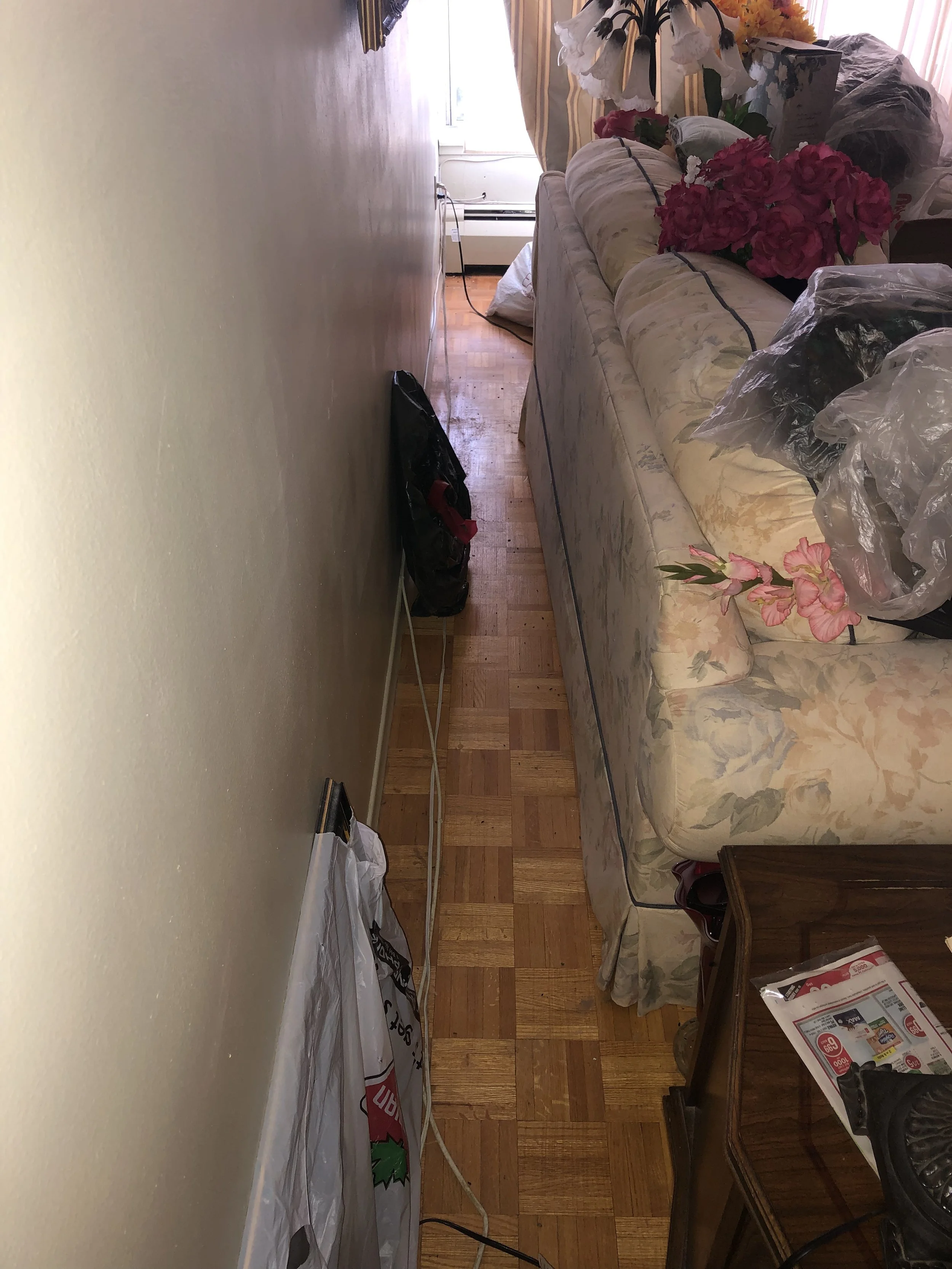 A living room with a floral sofa, various plastic bags, and a table with magazines. There are flowers on the sofa and a window with curtains in the background.