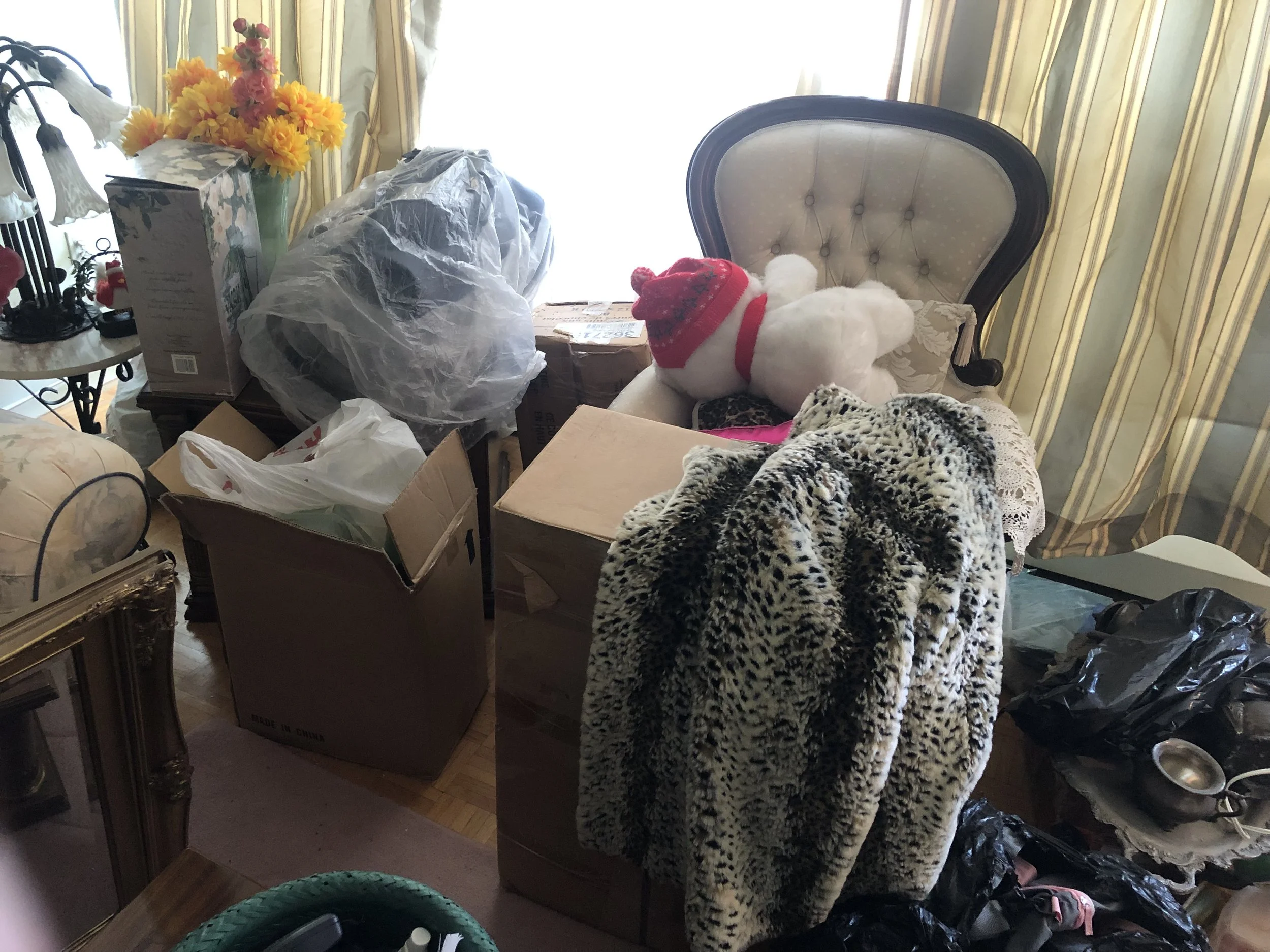 A cluttered room with various items including boxes, a teddy bear with a red hat, a leopard print blanket draped over a chair, and a vase with yellow and pink flowers near a window with yellow curtains.