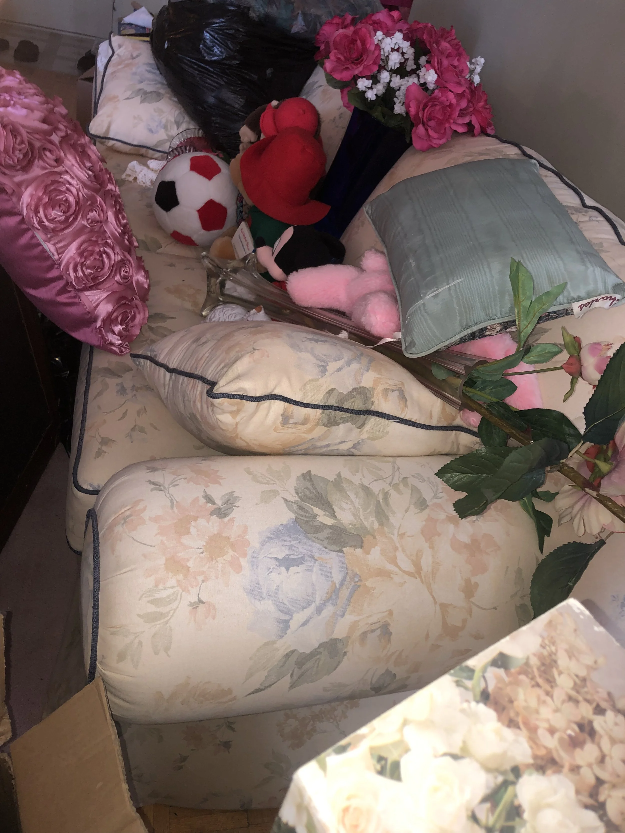 A floral-patterned sofa with various stuffed animals, a pillow, a flower arrangement, and some miscellaneous items on it.