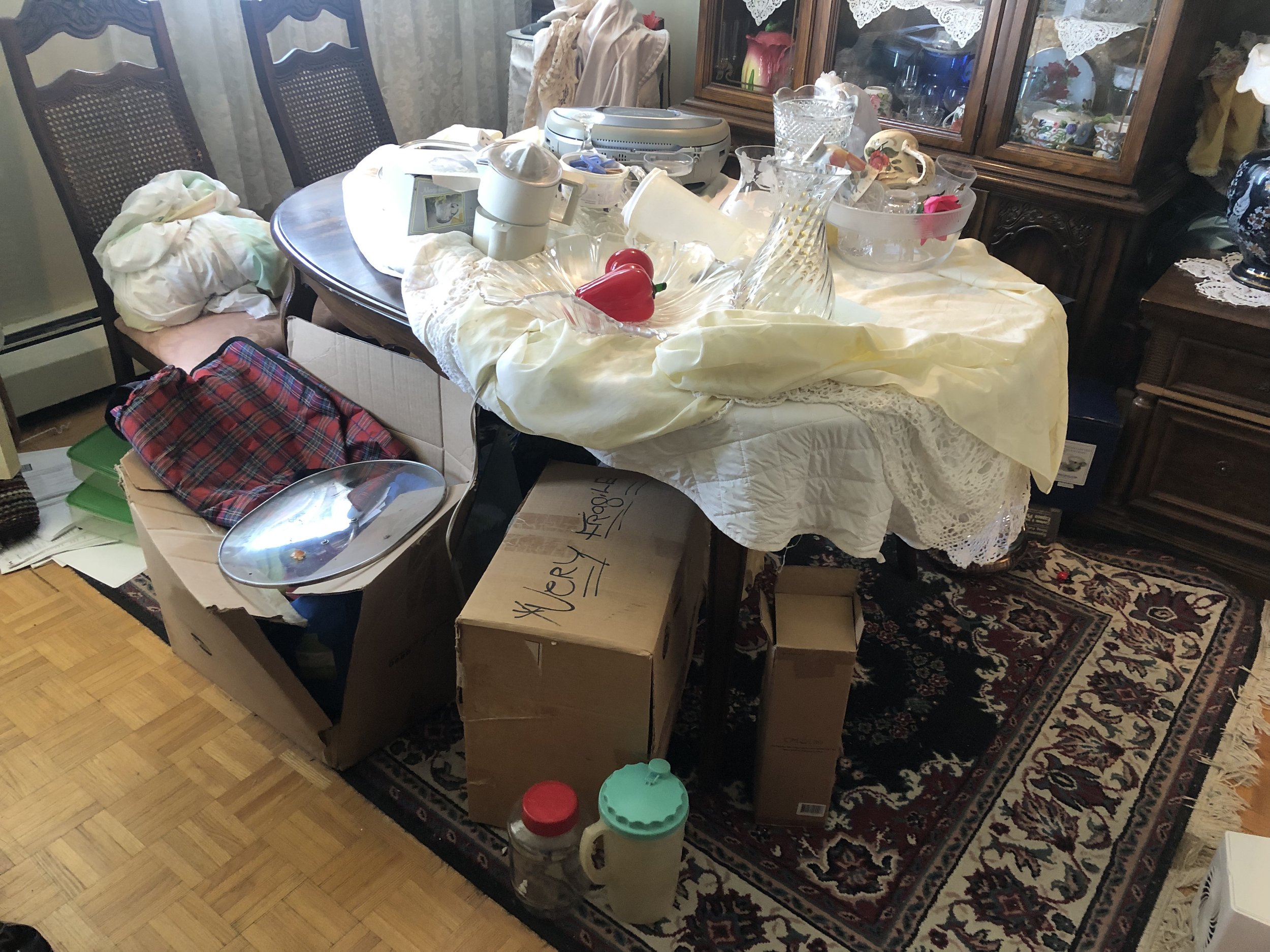 Cluttered dining room table with various household items, dishes, and cardboard boxes, surrounded by chairs and a wooden china cabinet.