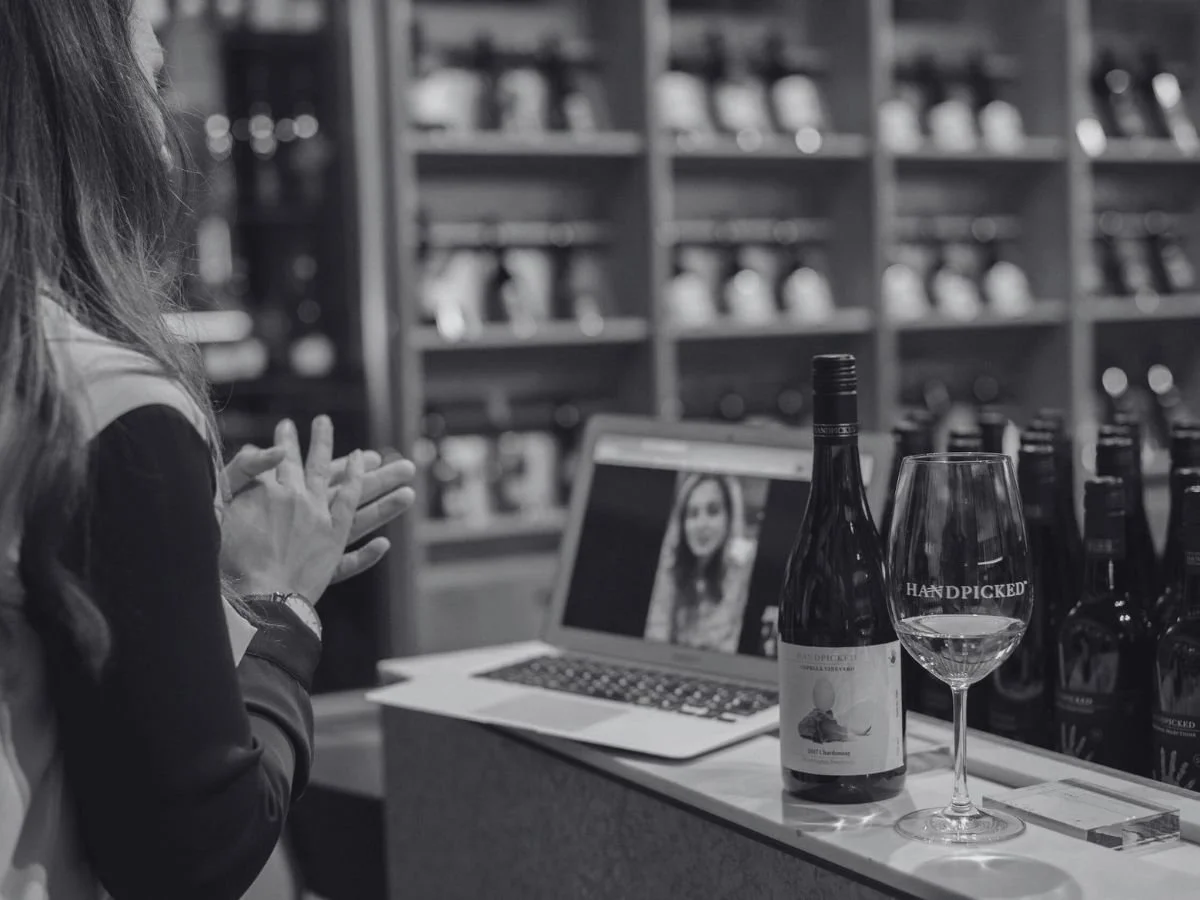 Woman with long hair sitting at a bar, on a video call on her laptop, with wine bottles and a glass of wine on the bar in front of her.