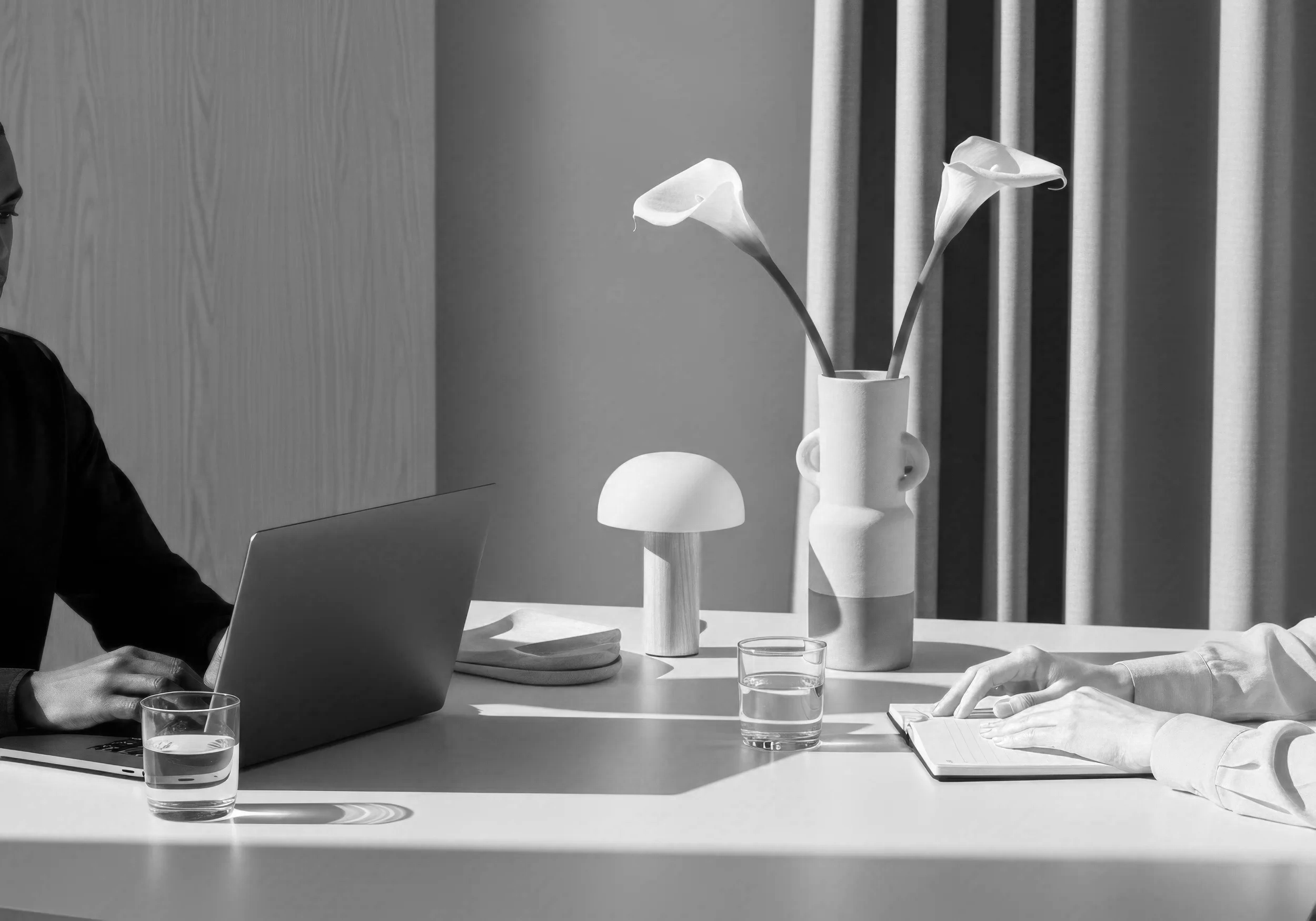 Two people sitting at a table. The left person is using a laptop and the right person is taking notes on a notepad. There are two glasses of water, a decorative vase with calla lilies, and a small lamp on the table. The setting appears modern with curtains in the background.