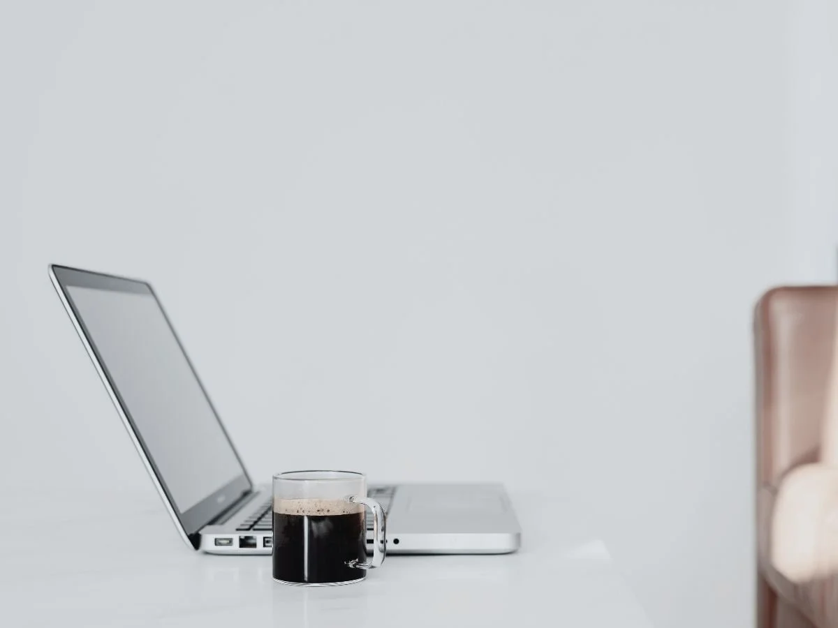 Open laptop and glass mug of black coffee on a minimalist white desk.