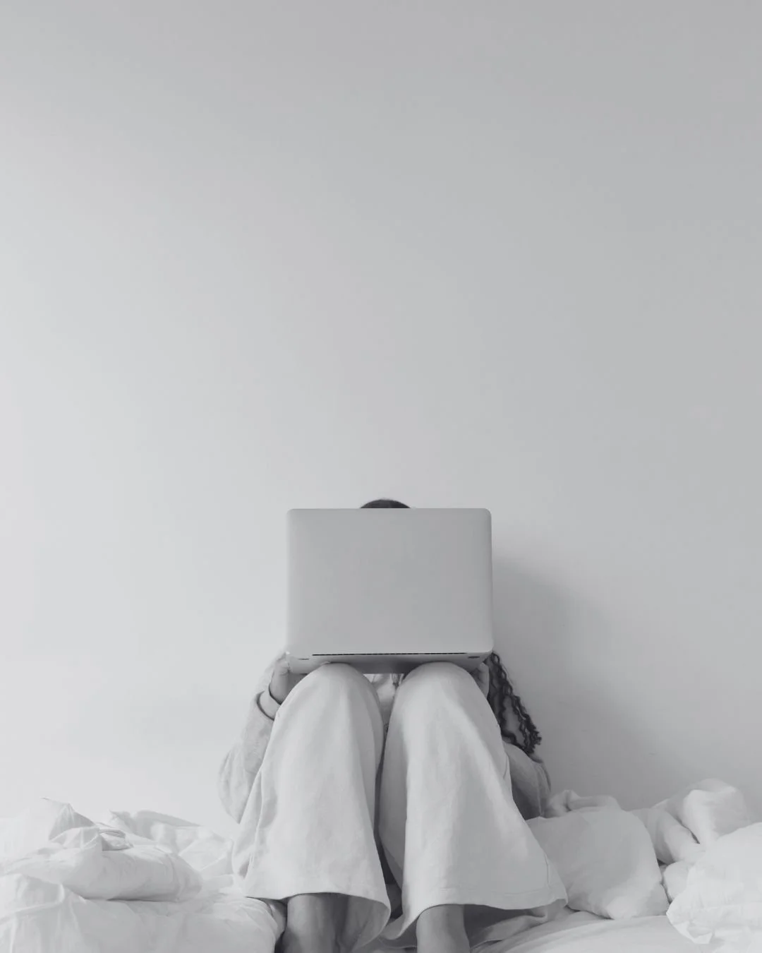 Person sitting on a bed with a laptop resting on their knees, working against a plain white wall.