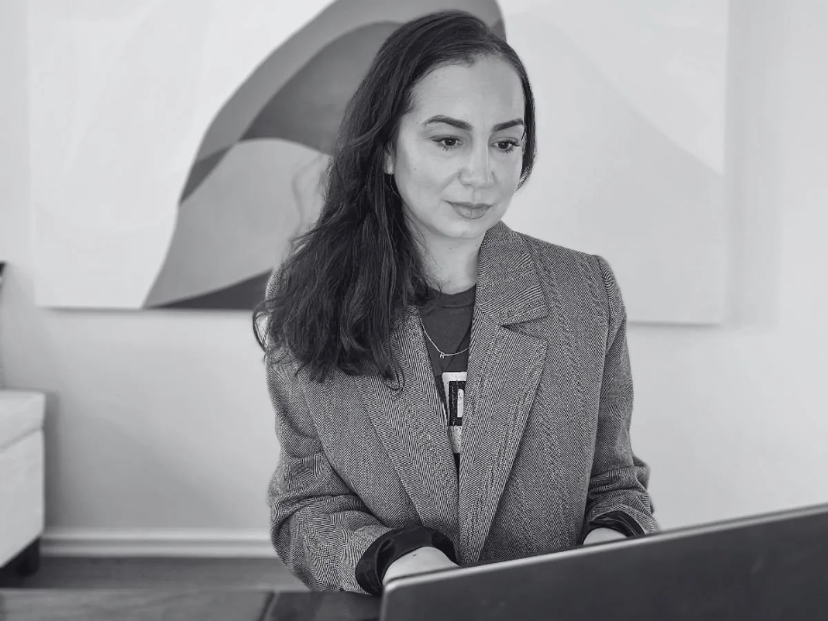 A woman with long dark hair looking at a laptop screen, dressed in a blazer and T-shirt, in an indoor setting with modern art on the wall behind her.