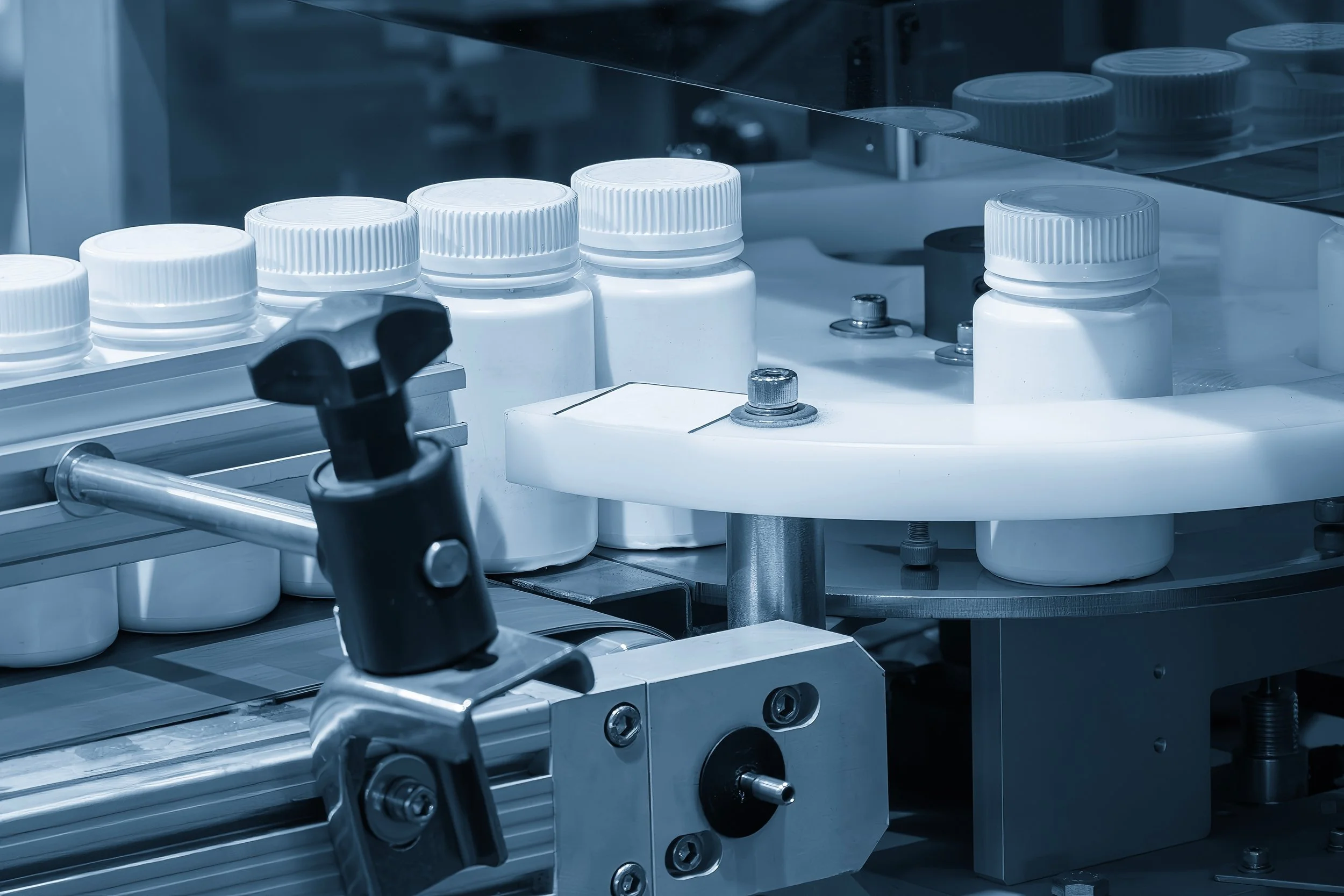 Close-up of a pharmaceutical production line with several white pill bottles and industrial machinery.