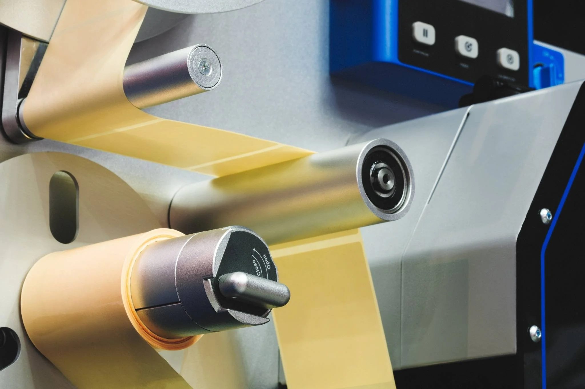 Close-up of a medical or industrial device with metallic rollers and a yellow sheet being processed. The device includes knobs and controls on a metallic panel.