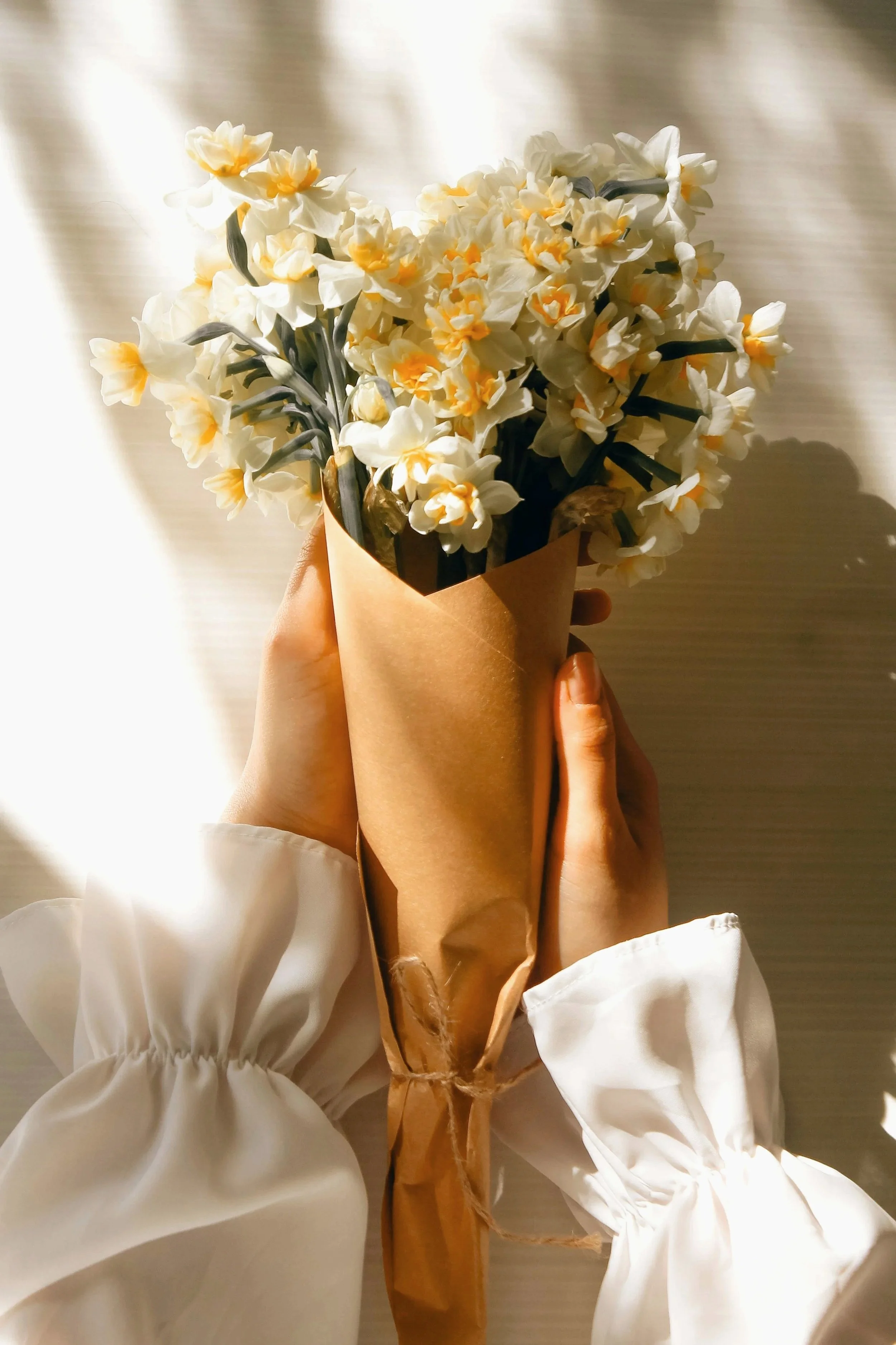 A person holding a bouquet of white and yellow flowers wrapped in brown paper, with sunlight shining on it.