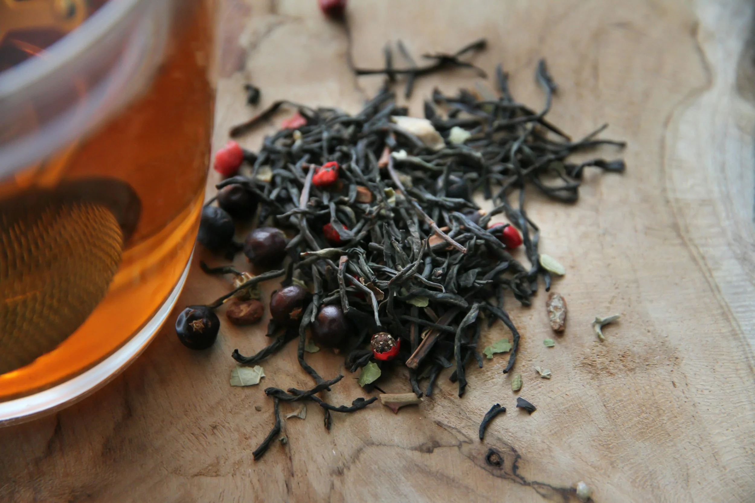 Dried black tea leaves with some red and purple berries and loose green herbs on a wooden surface, next to a glass of brewed tea.