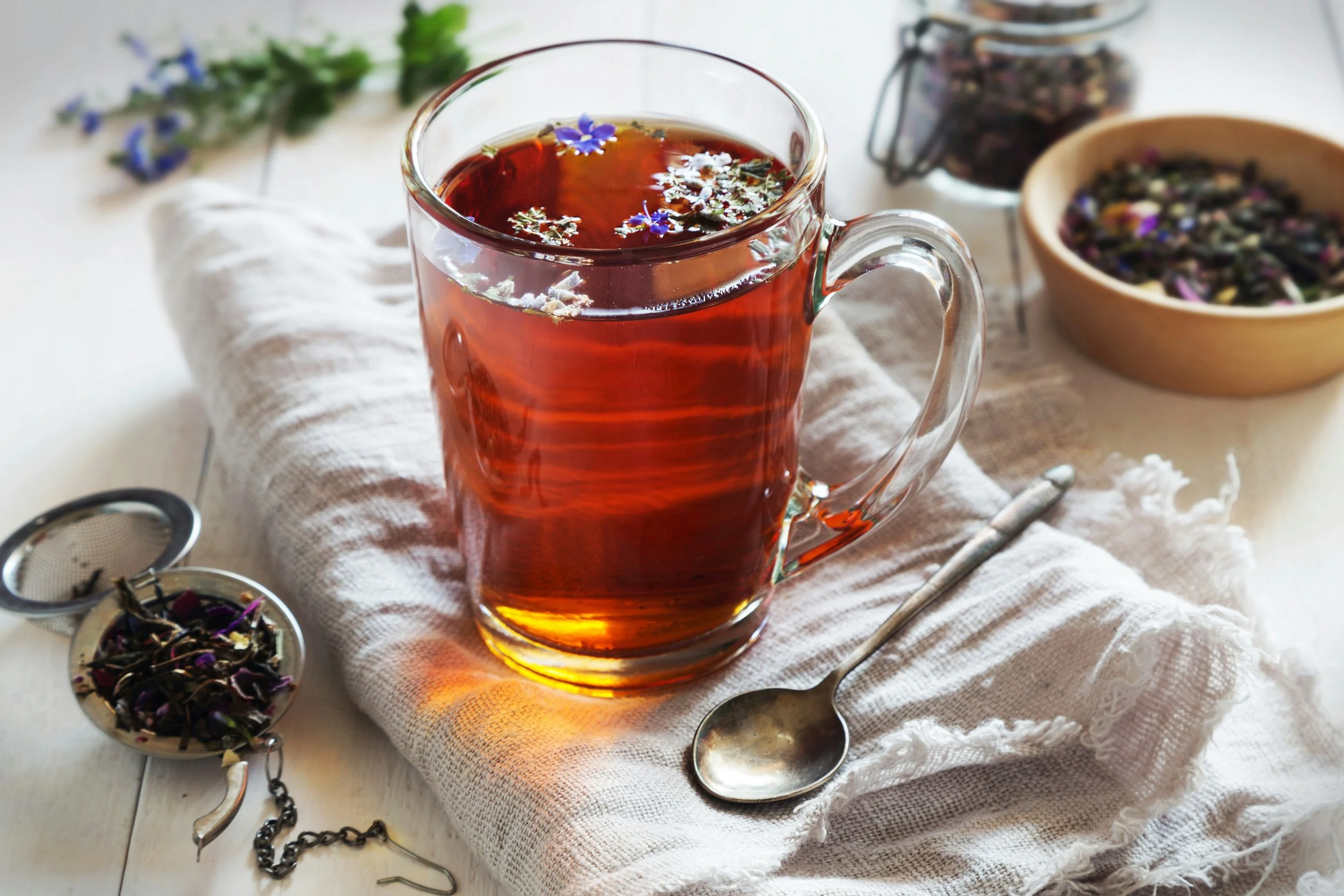 A glass mug of herbal tea with blue and white flowers floating on top, placed on a white cloth napkin, with loose tea leaves and a teaspoon nearby, on a light-colored surface.